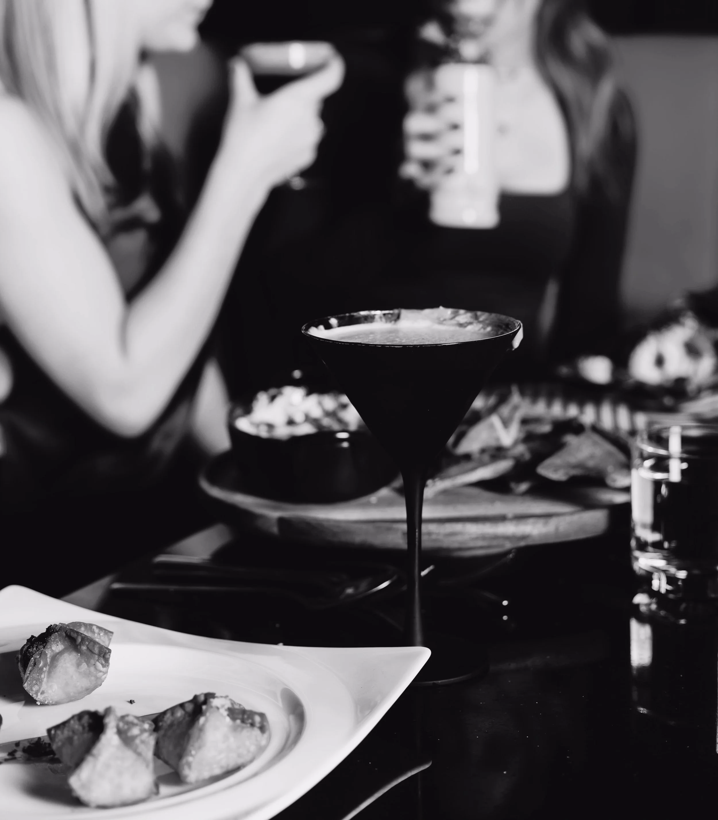 A sleek black martini glass surrounded by food on a table, showcasing the elevated cocktail and dining experience at Silk Utica cocktail and dessert bar in Utica, New York.