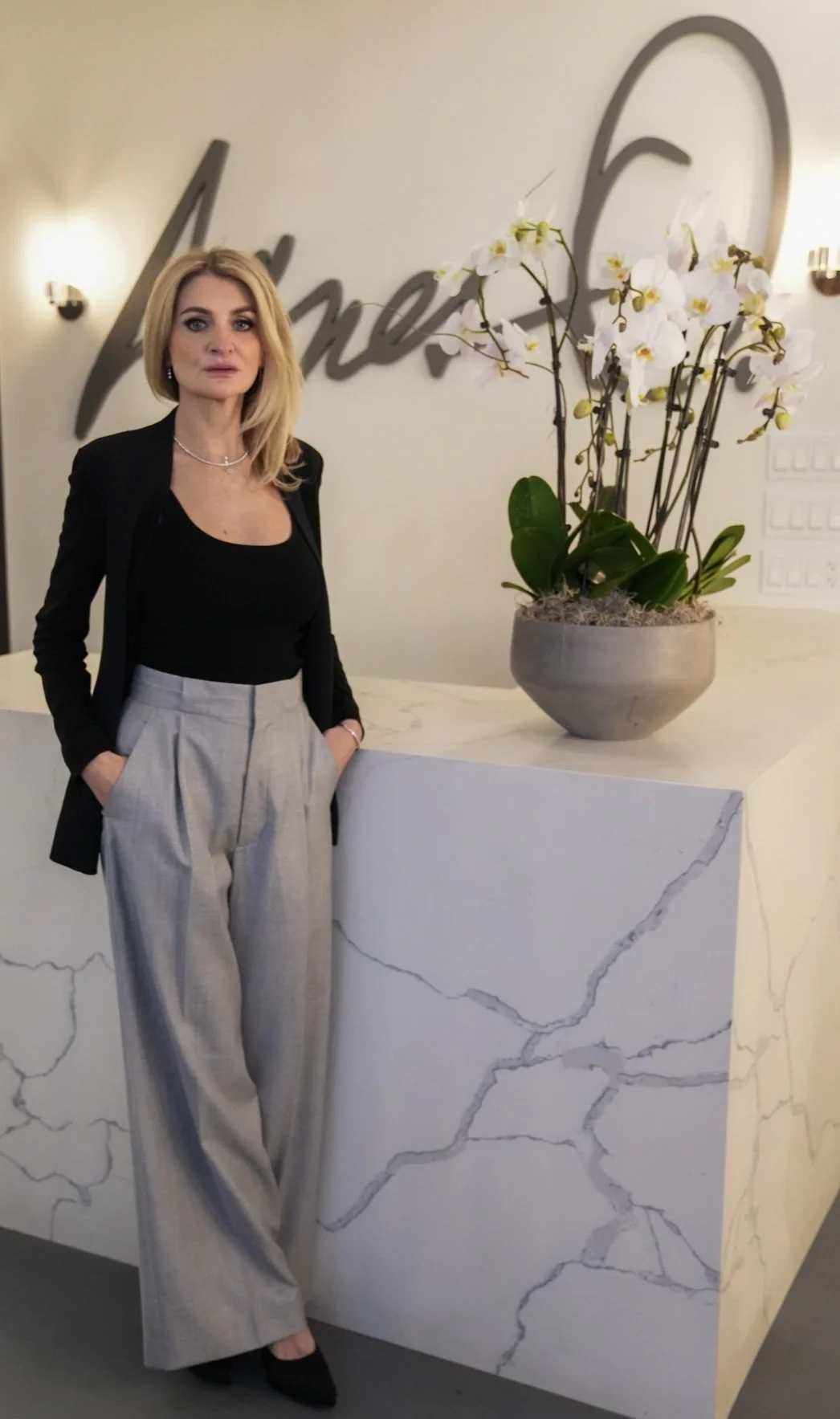A woman with blonde hair wearing a black top, black blazer, and gray plaid pants standing beside a white marble counter with dark veining and a large orchid plant in a gray pot. There is a sign on the wall behind her with writing, and she is posed with her hands in her pockets.