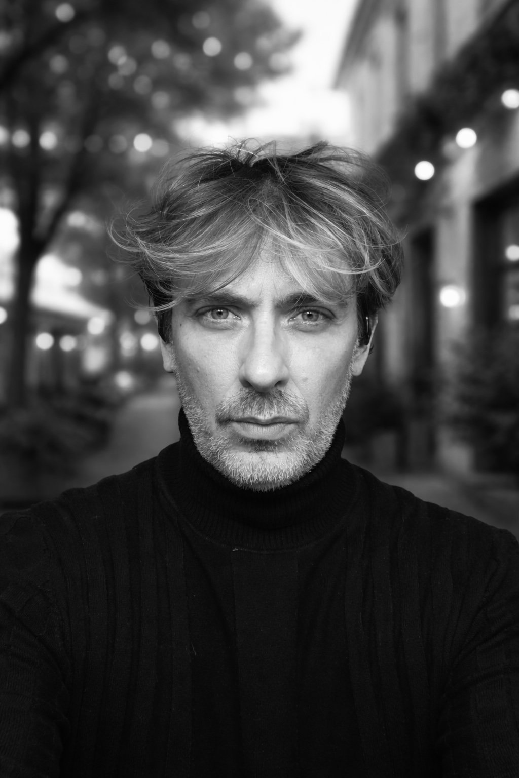 Black and white portrait of a man with tousled hair and a beard, wearing a black turtleneck, outdoors at night with blurred lights and trees in the background.