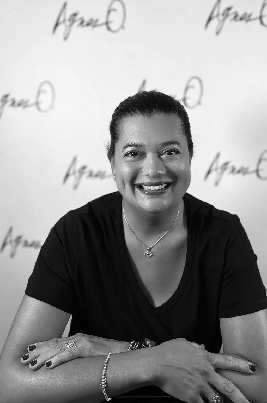 Black and white portrait of a smiling woman with shoulder-length hair, wearing a dark blouse and a necklace, against a plain light gray background.