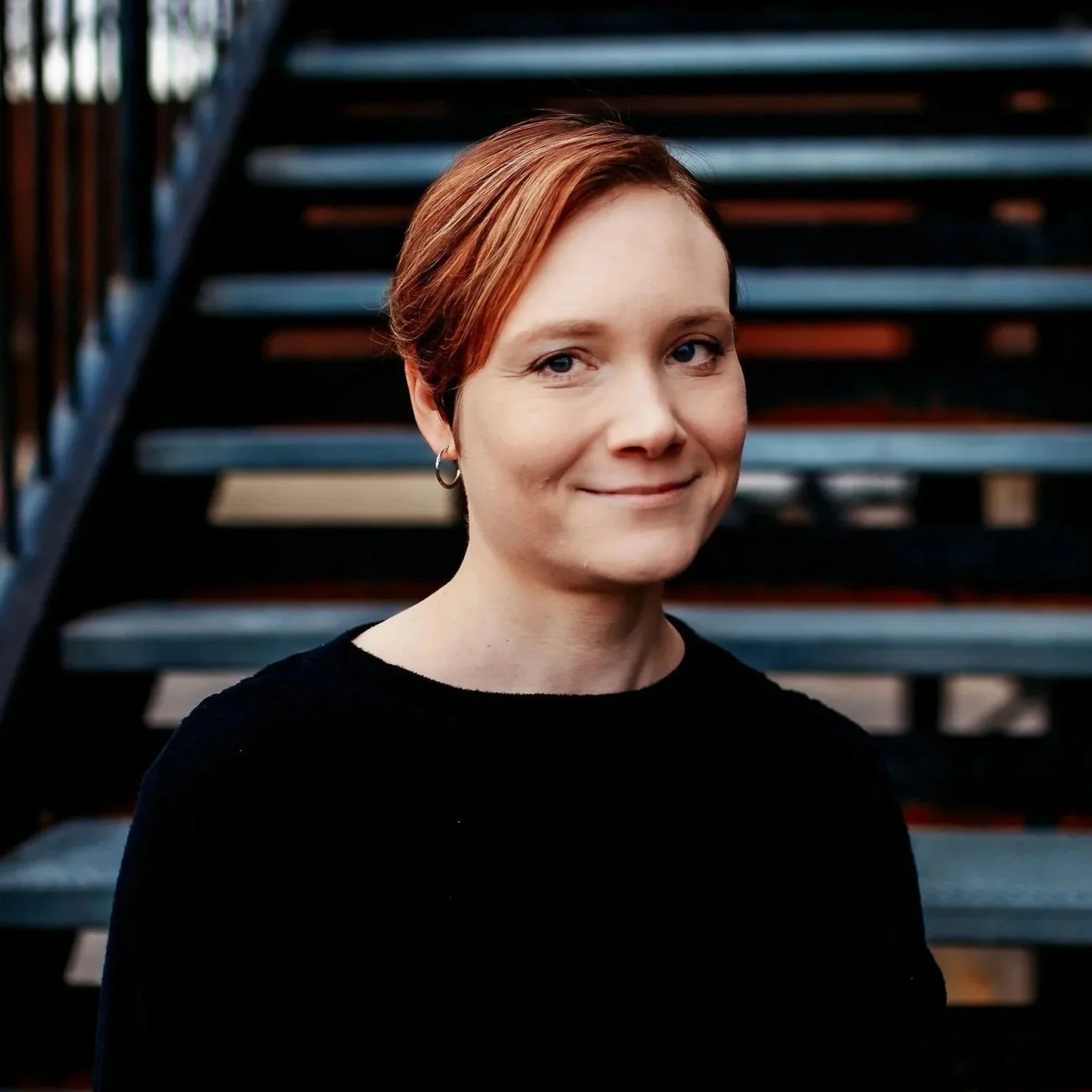 Occupational Therapist Lauren Kapolnek, a woman with short reddish hair, wearing a black top and grey jeans, sitting on metal stairs outdoors.
