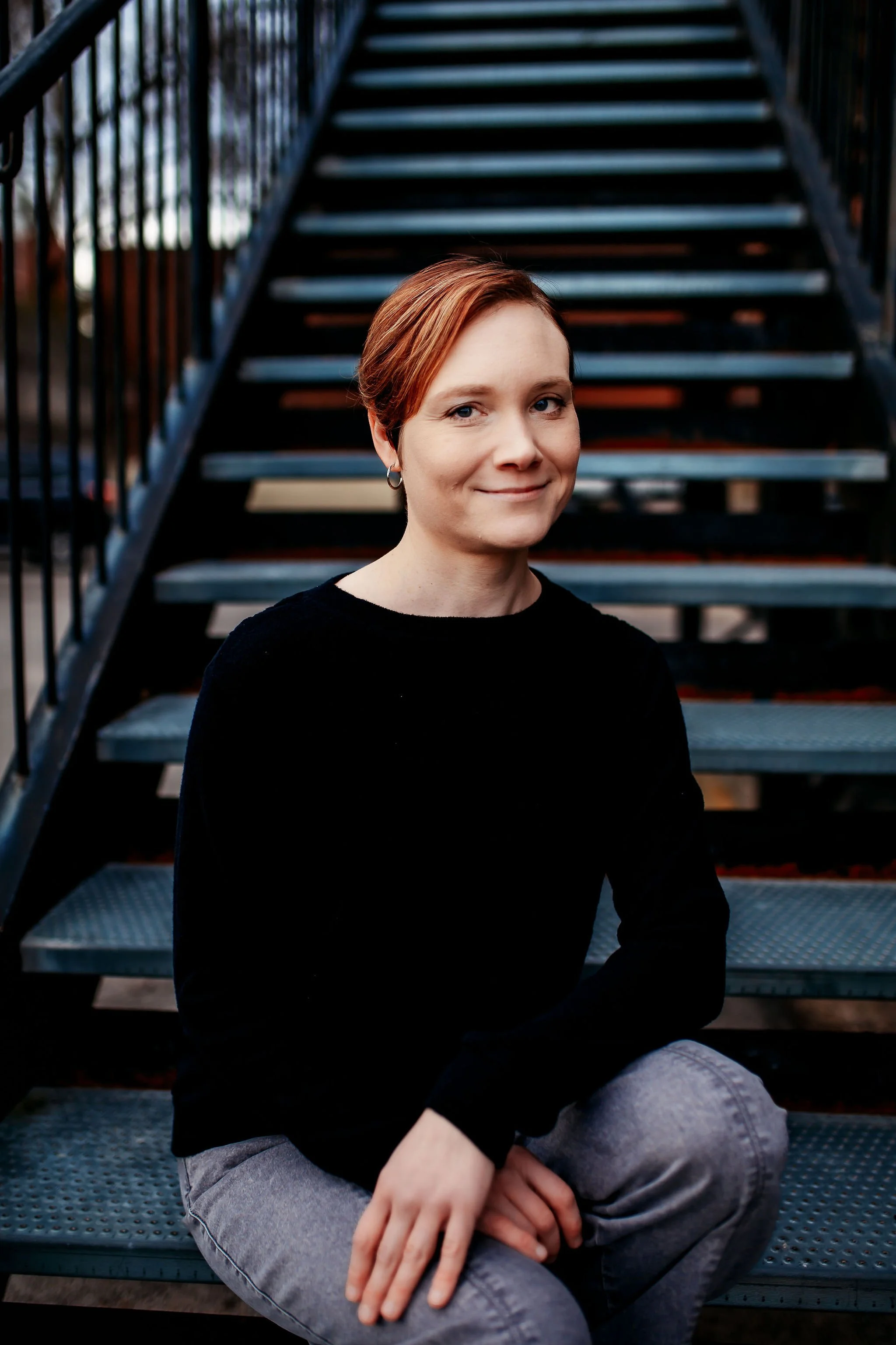 A woman with short red hair and hoop earrings sitting on metal stairs outdoors, wearing a black top and gray pants.