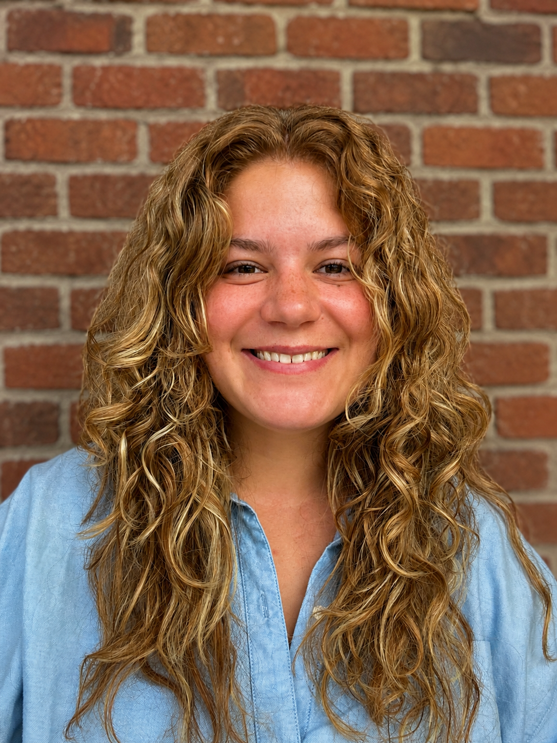 Therapist Sarah Newell with long curly hair smiling in front of a brick wall wearing a light blue button-up shirt.
