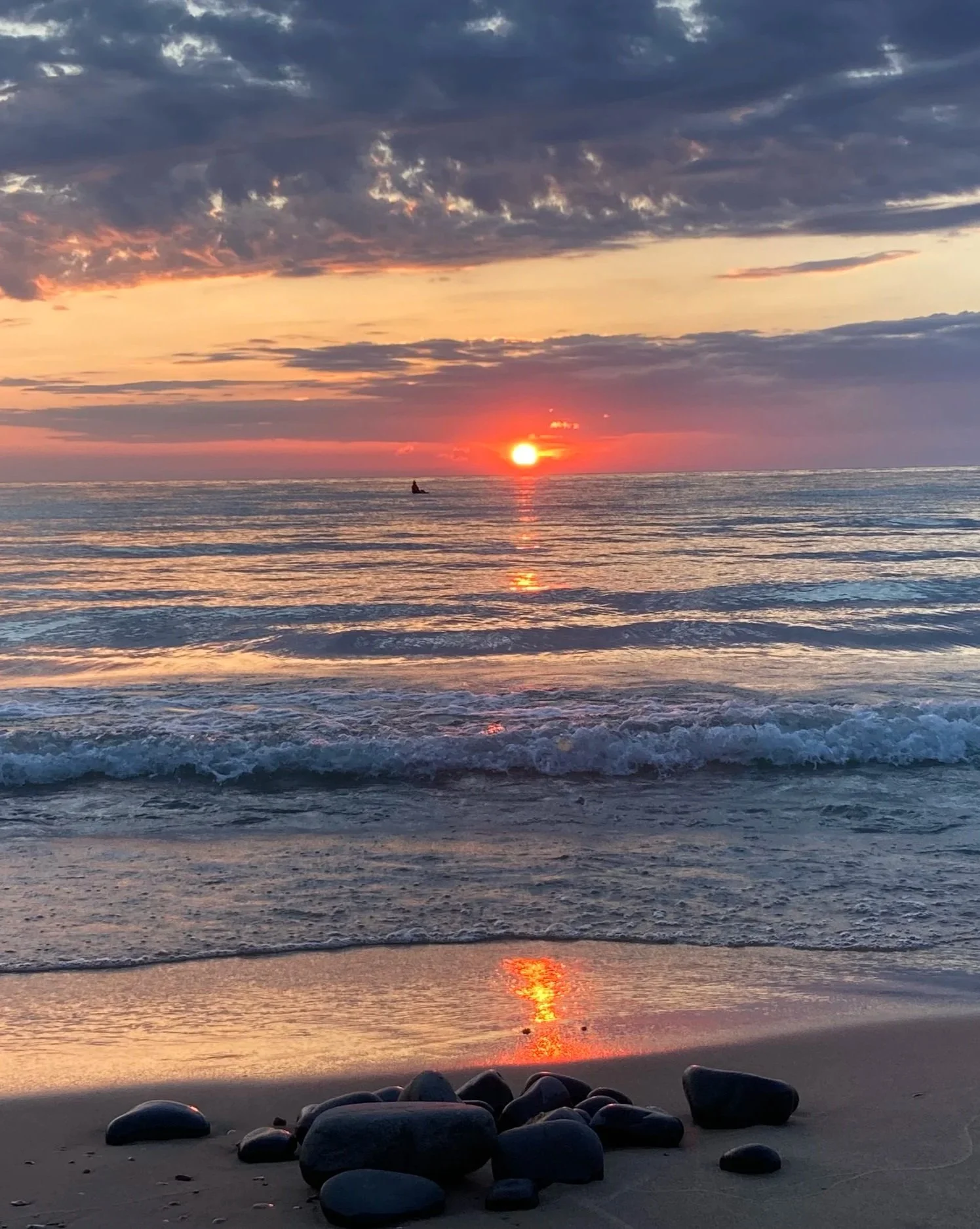 Sunset over Lake Michigan with a small boat in the distance, waves lapping against the shore with smooth rocks in the foreground.