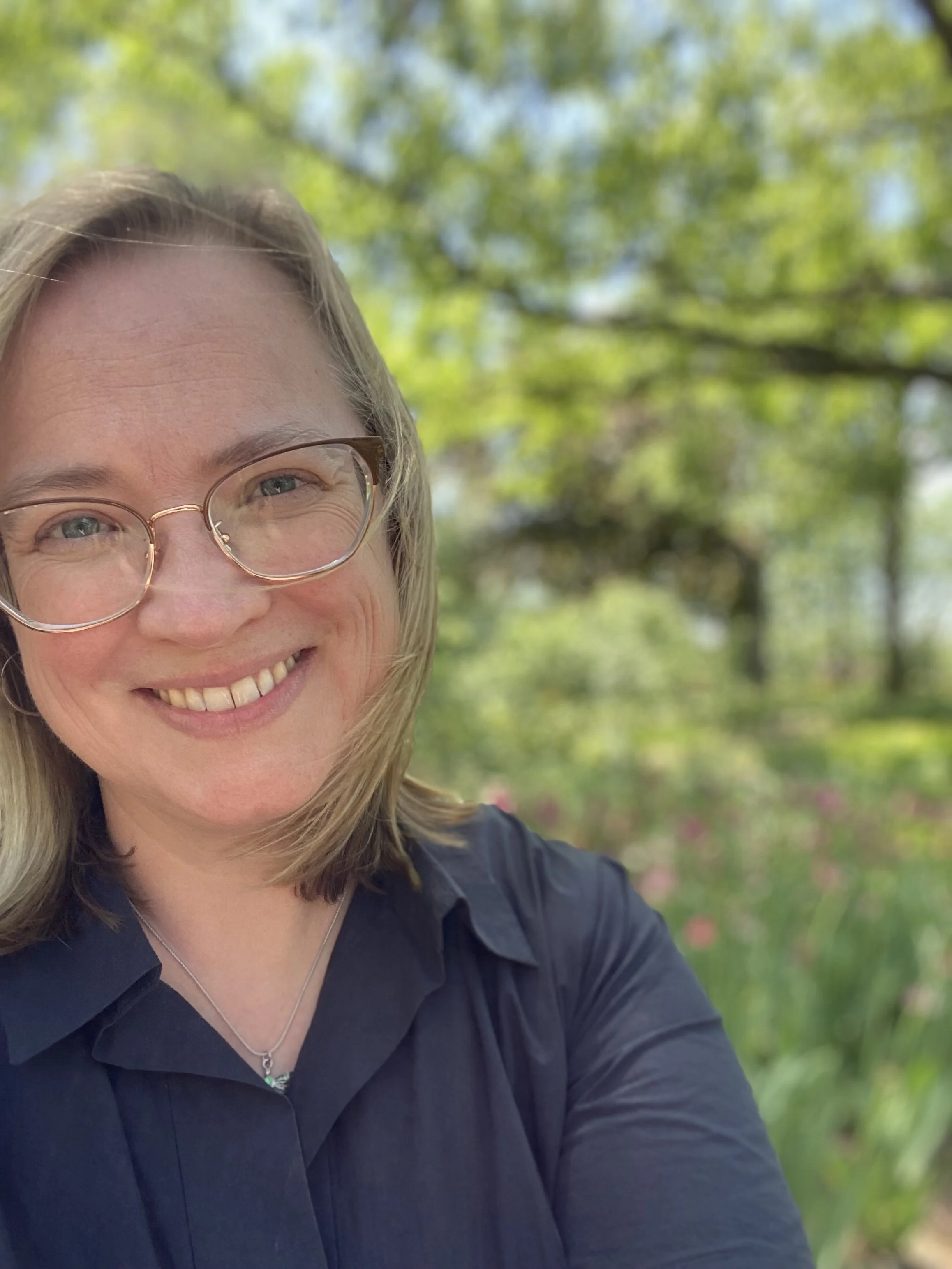 Close up of Therapist Ruth Farrell with blonde hair and glasses outdoors, with green trees and sunlight in the background.