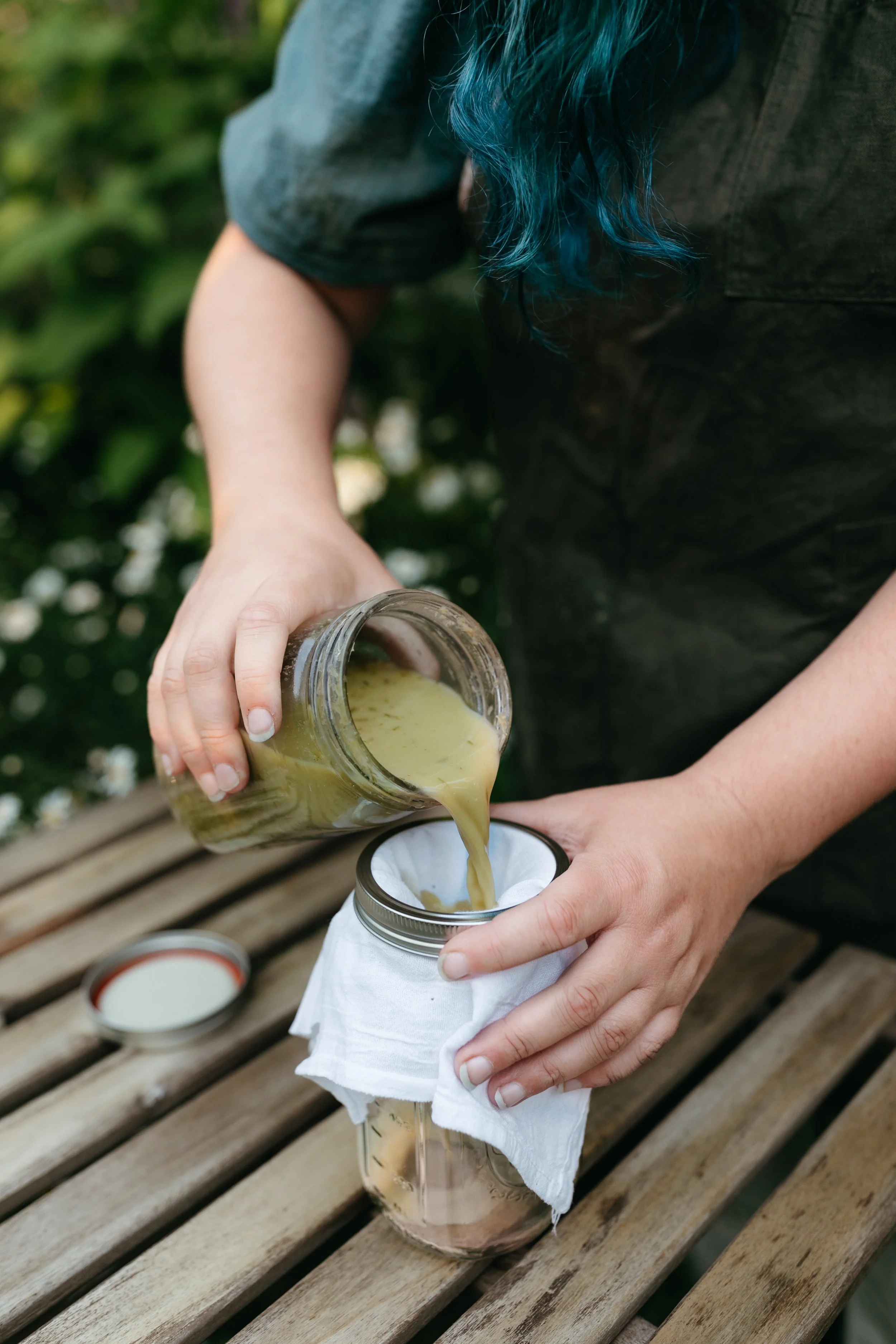 Person pouring green liquid from a jar through a cloth filter into another jar on a wooden table.