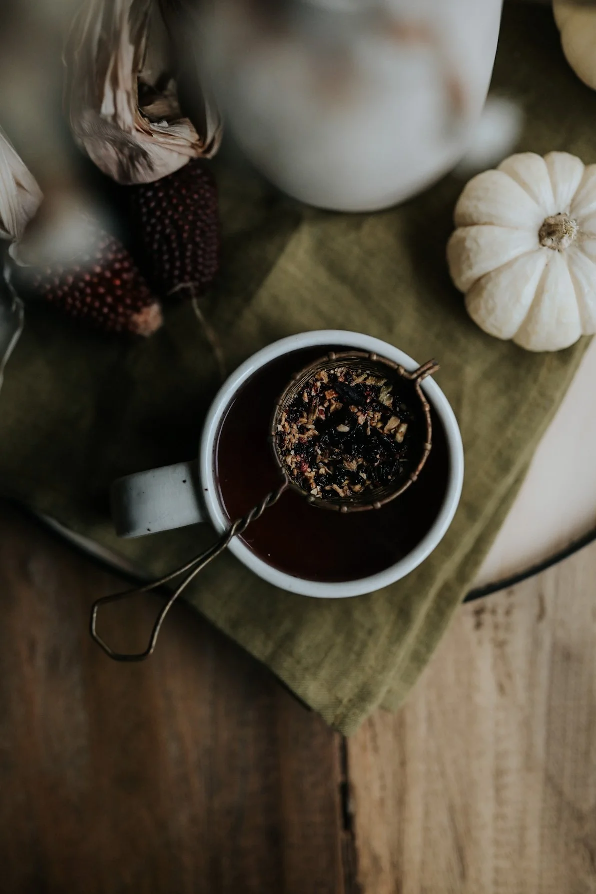 Overhead view of a cup of tea with a tea strainer, surrounded by decorative corn and a white pumpkin on a wooden surface with a green cloth.