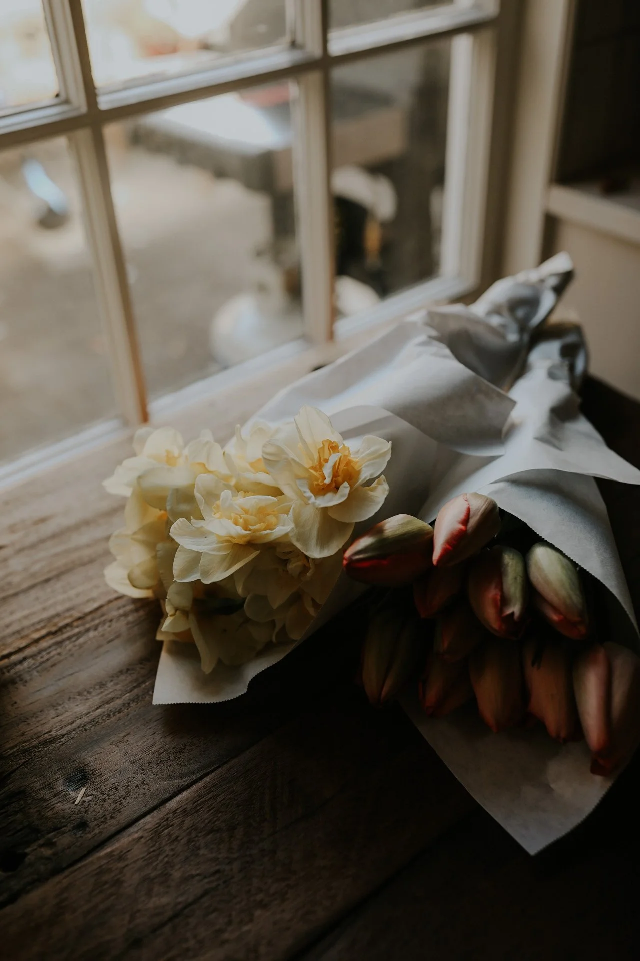 Bouquets of cream daffodils and tulips wrapped in paper on a wooden table by a window.