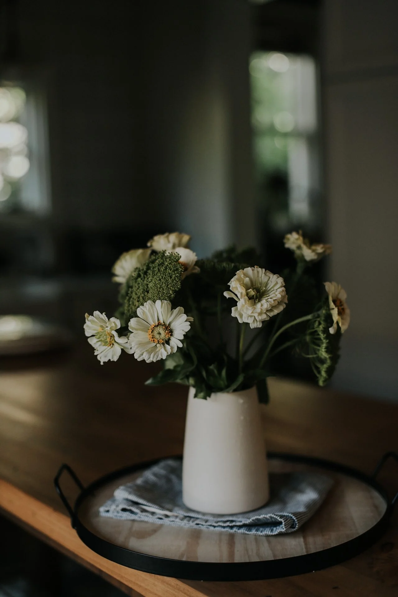White flowers in a white vase on a wooden table with a cloth on a tray, dimly lit room