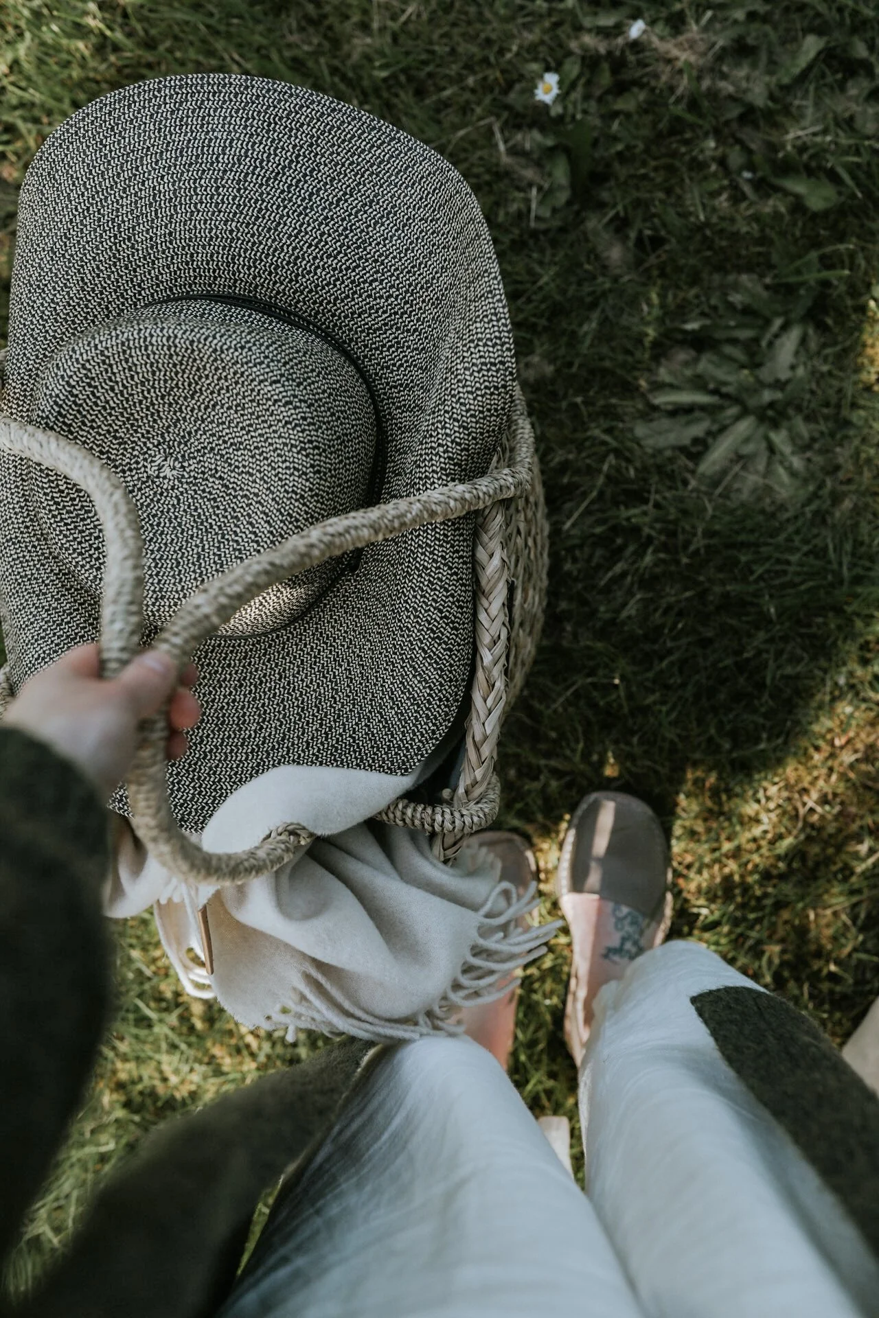 Top view of a person holding a woven bag with a textured hat and cream-colored scarf inside, standing on grass with visible shoes.