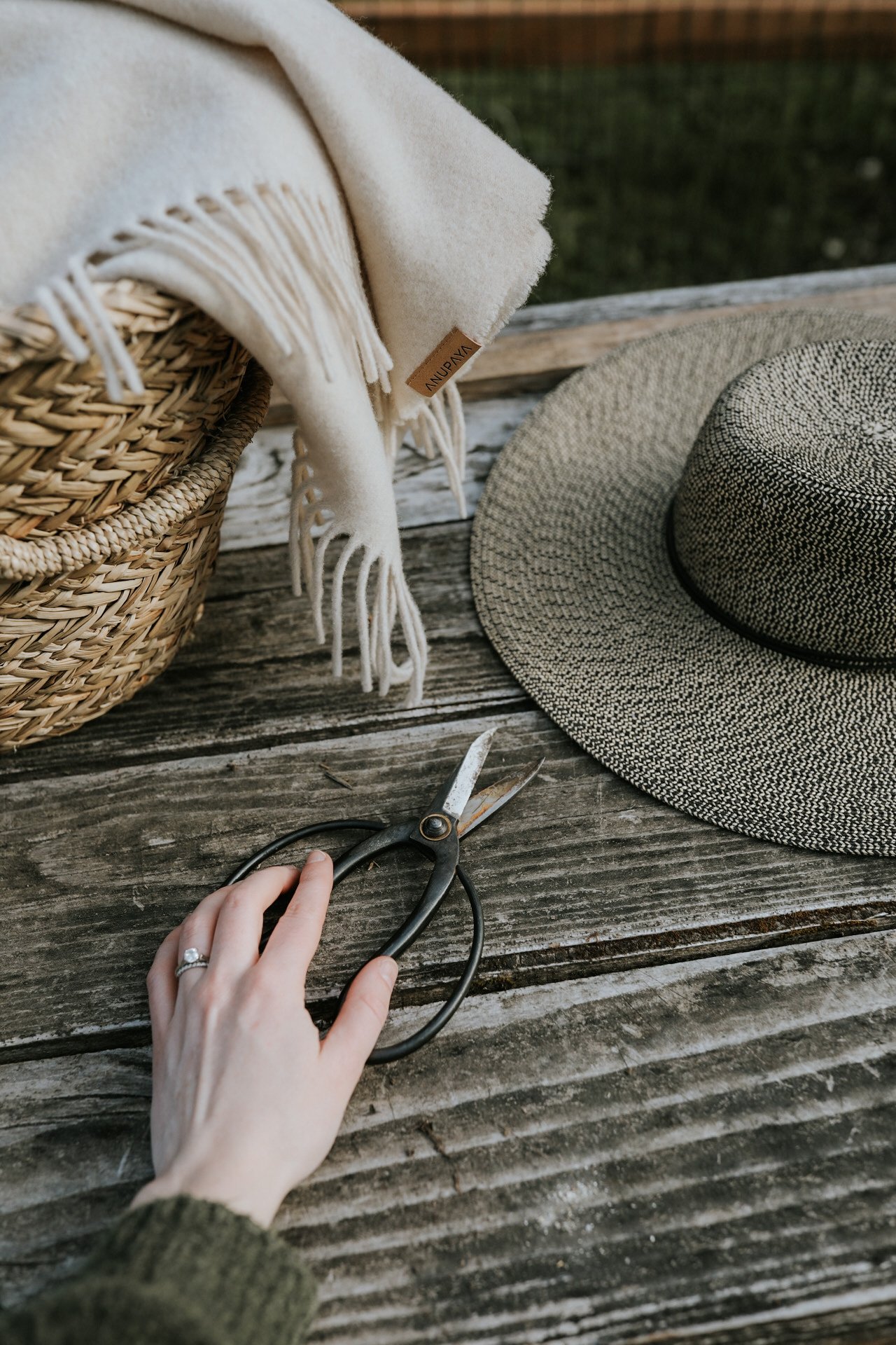 Wicker basket with white blanket, wide-brimmed hat, hand holding pruning shears on wooden table.
