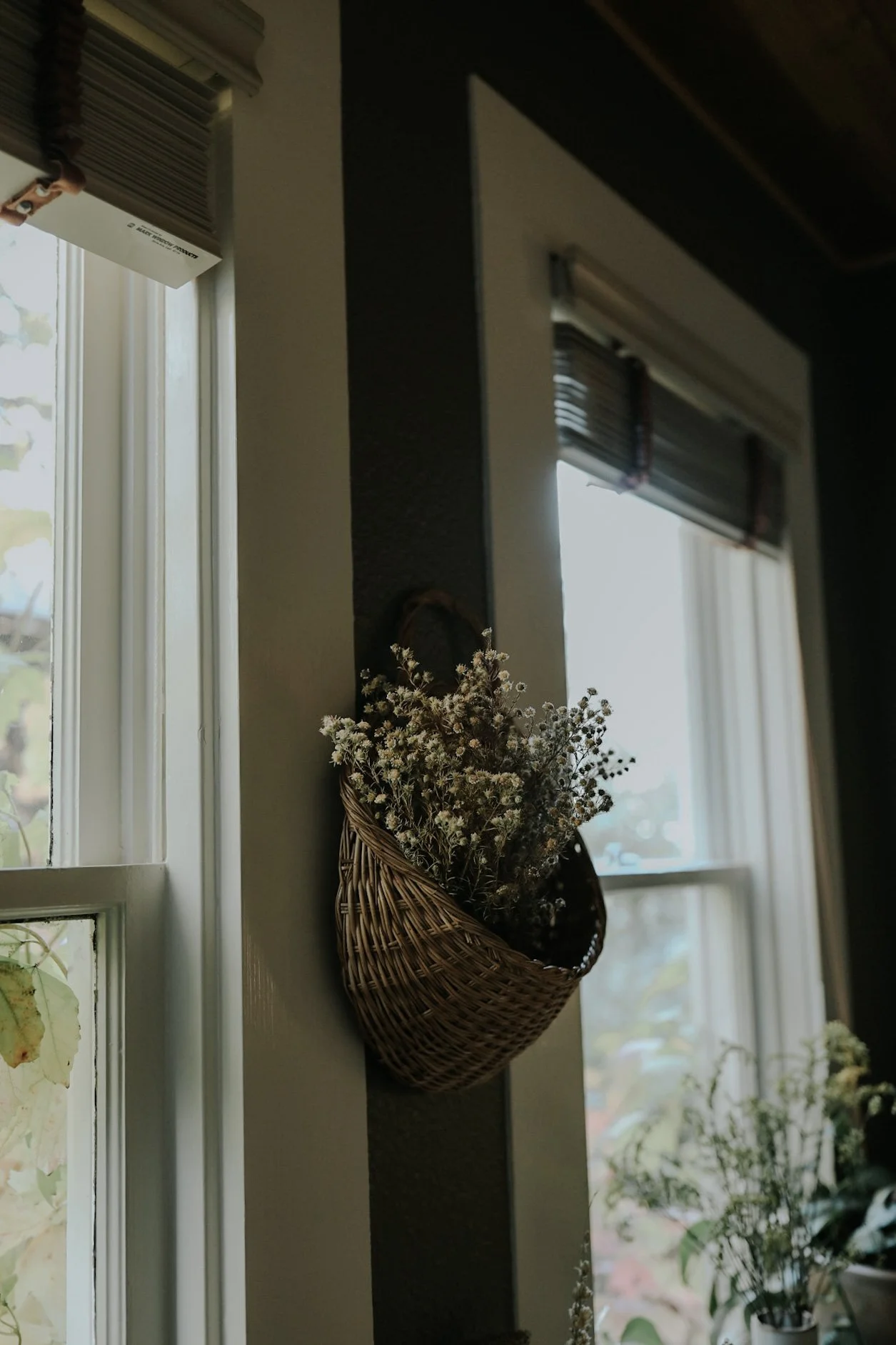 Wicker basket with dried flowers hanging on wall near window with blinds.