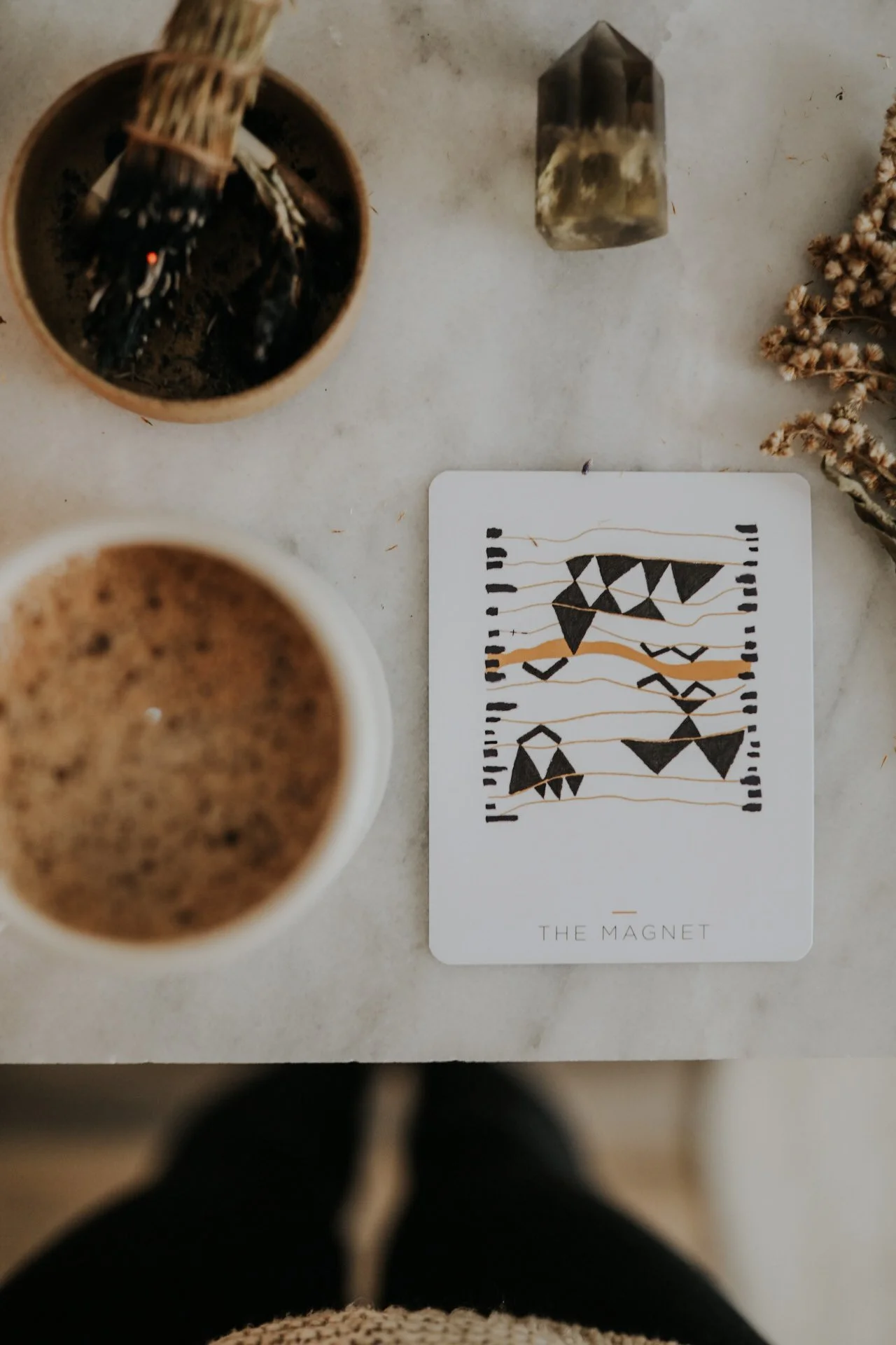 Marble countertop with a card labeled 'The Magnet,' crystal, sage bundle, coffee cup, and dried flowers.