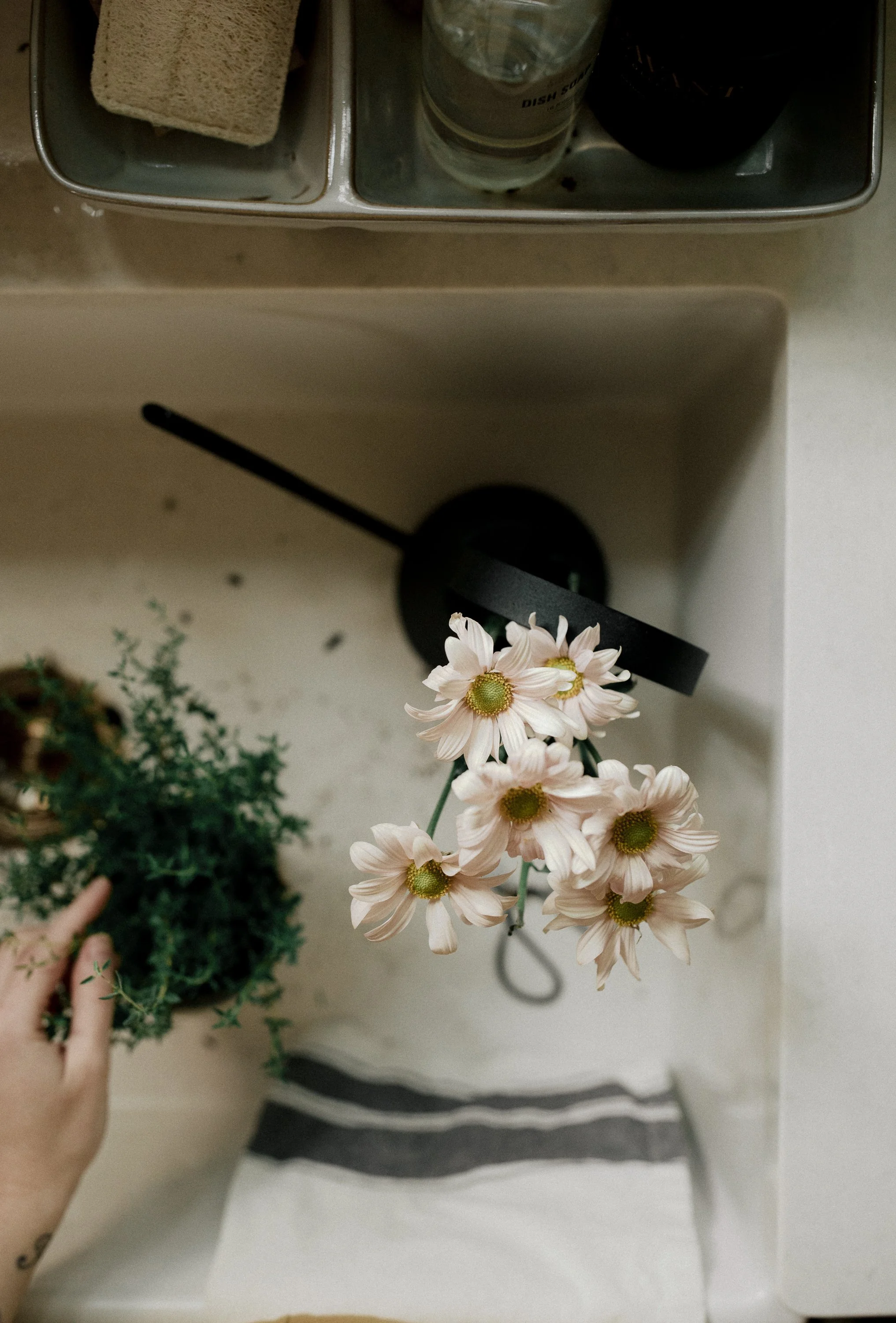 Top view of a kitchen sink with a bouquet of flowers, a hand holding a plant, dish soap, and a cloth.