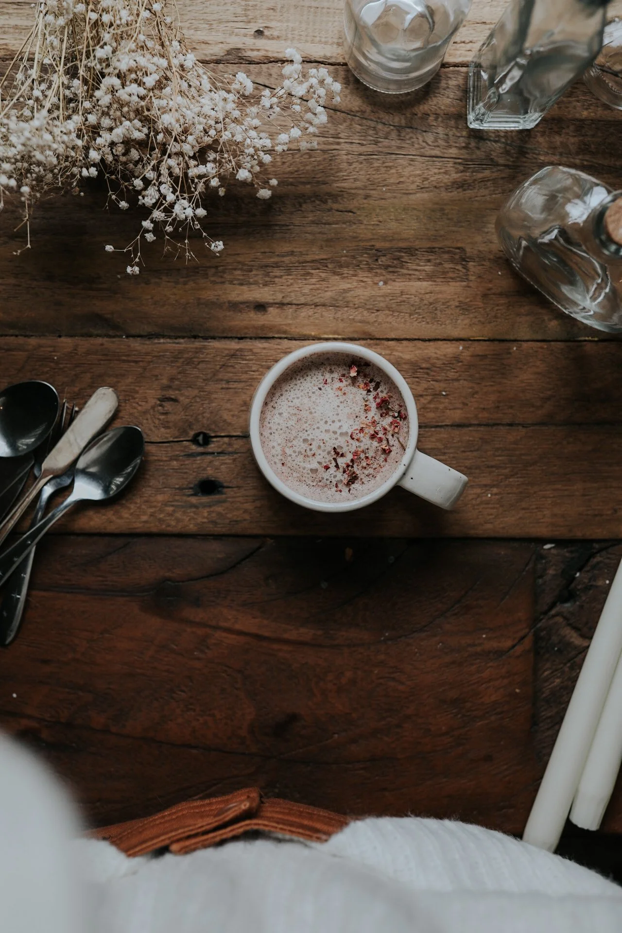 Cup of coffee with spices on wooden table, surrounded by spoons, glass bottles, and dried flowers.