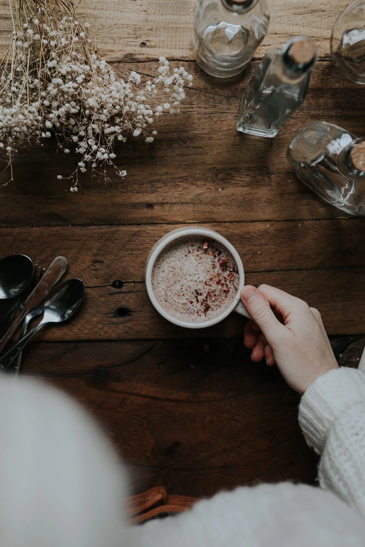 Hand holding a cup of cappuccino with cocoa powder on a wooden table, surrounded by dried flowers, spoons, and glass bottles.