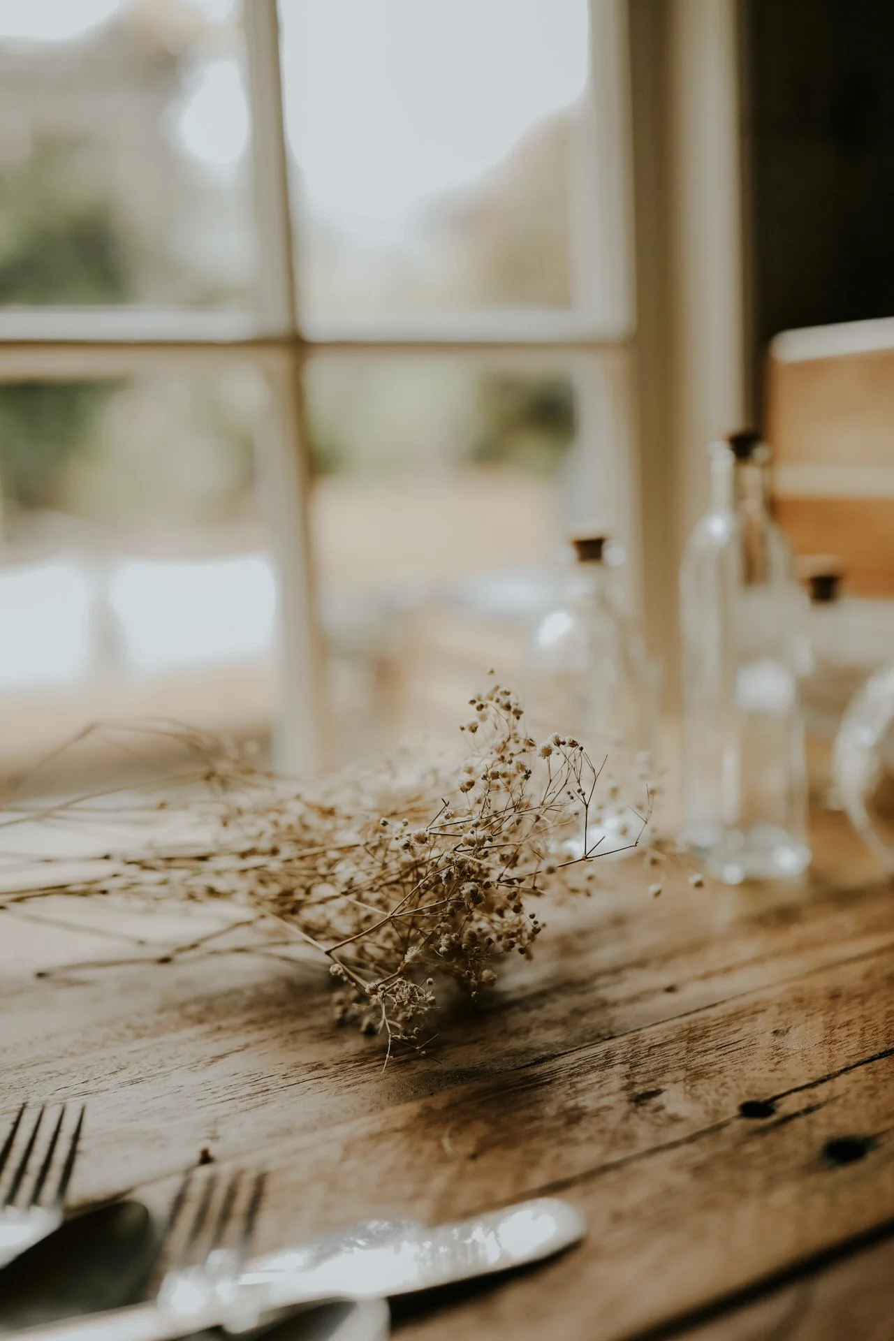 Rustic table setting with dried flowers and empty glass bottles in front of a window.