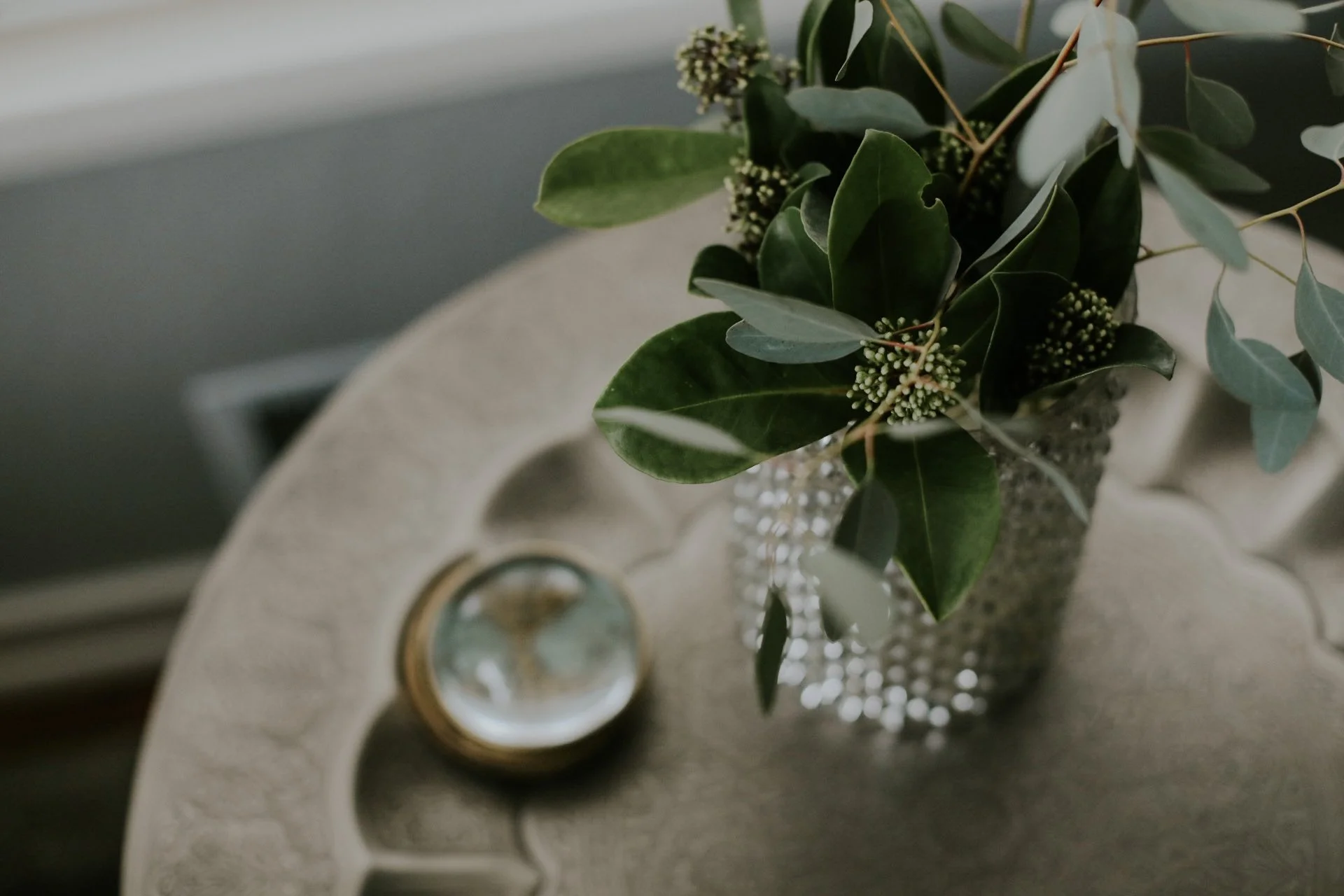 Decorative table with vase of green leaves and small plant, next to a magnifying glass in a brass holder