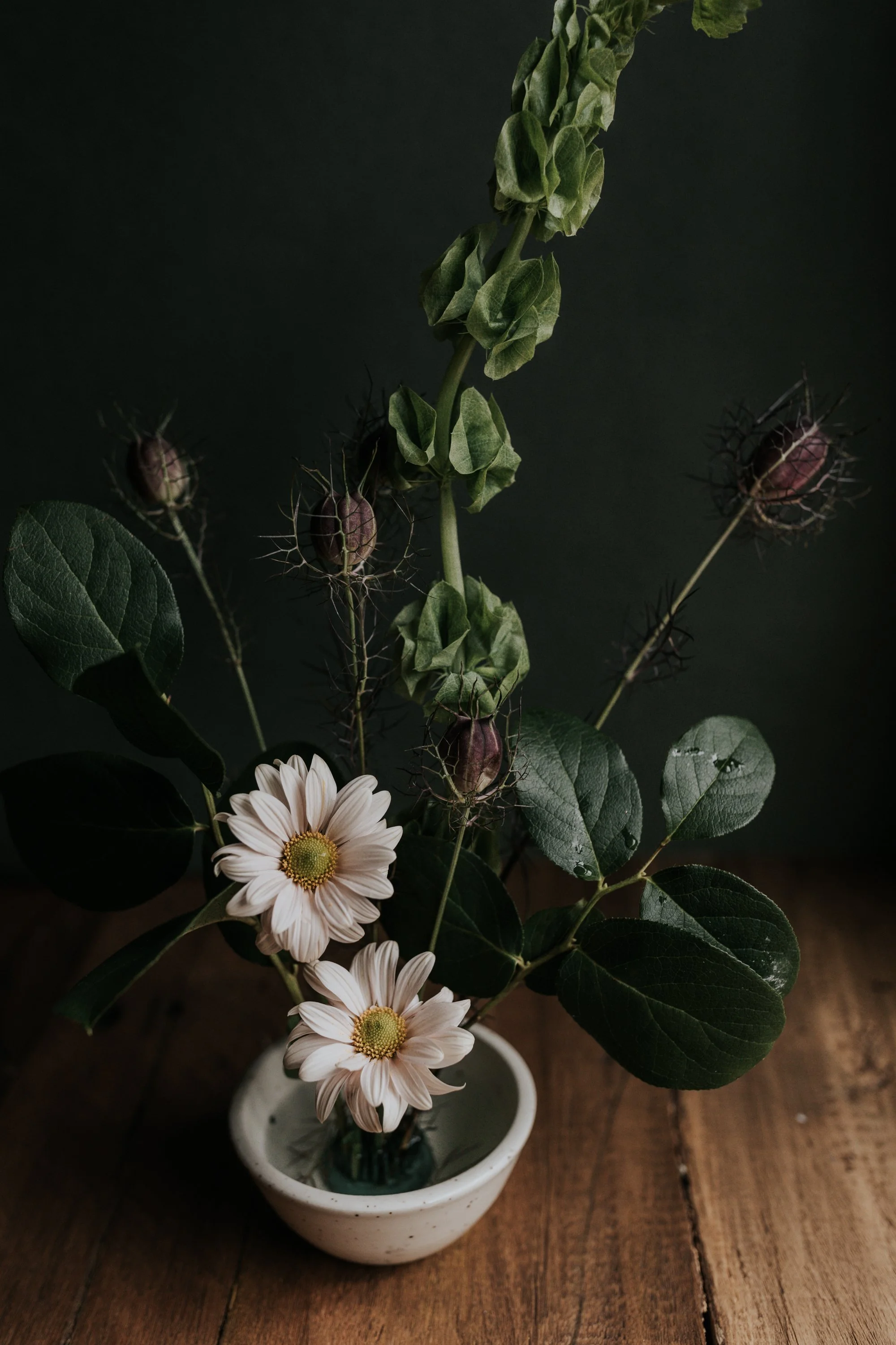 Rustic floral arrangement in a small ceramic bowl with white daisies, greenery, and dark seed pods on a wooden table.