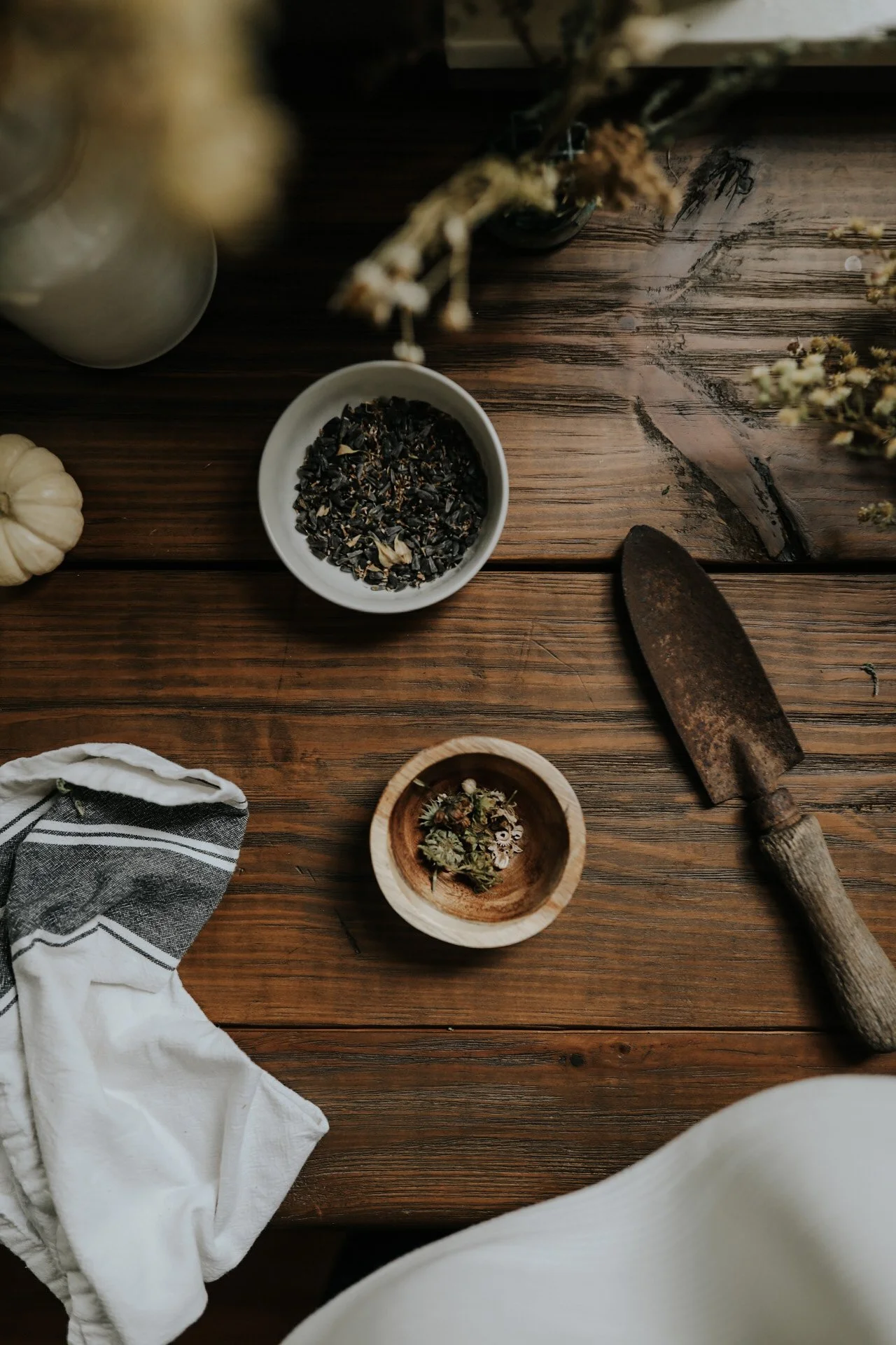 Rustic wooden table with dried herbs in bowls, a garden trowel, striped cloth, and a small white pumpkin.