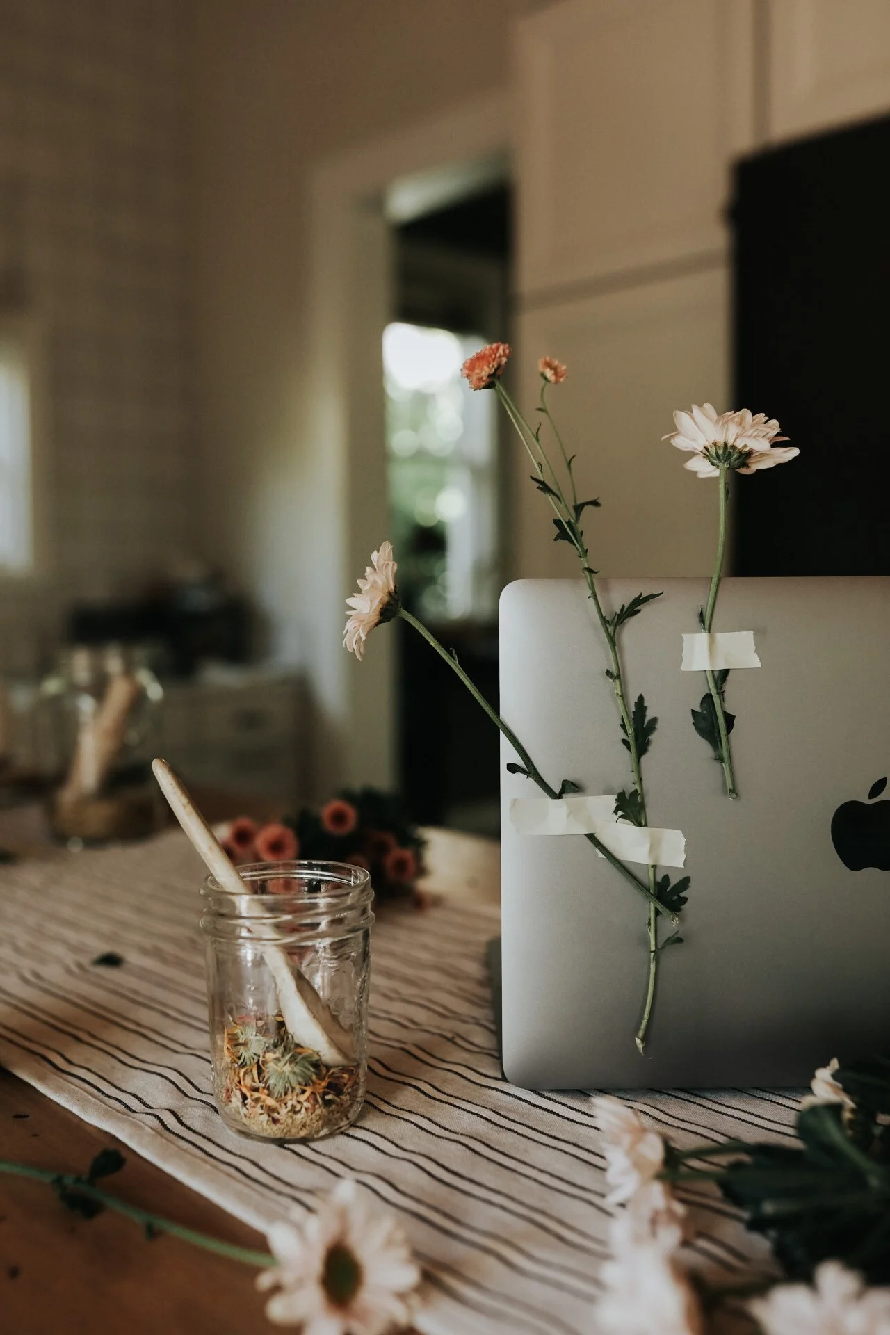 Open laptop with flowers taped to it, glass jar with dried flowers on a striped tablecloth, and blurred background.