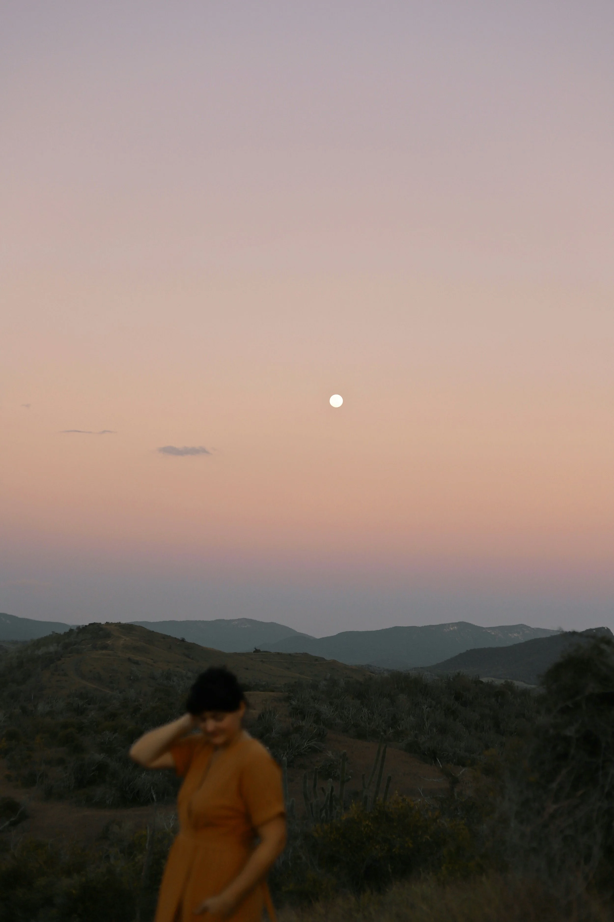 Blurry woman in orange dress with sunset landscape, mountains, and a full moon.