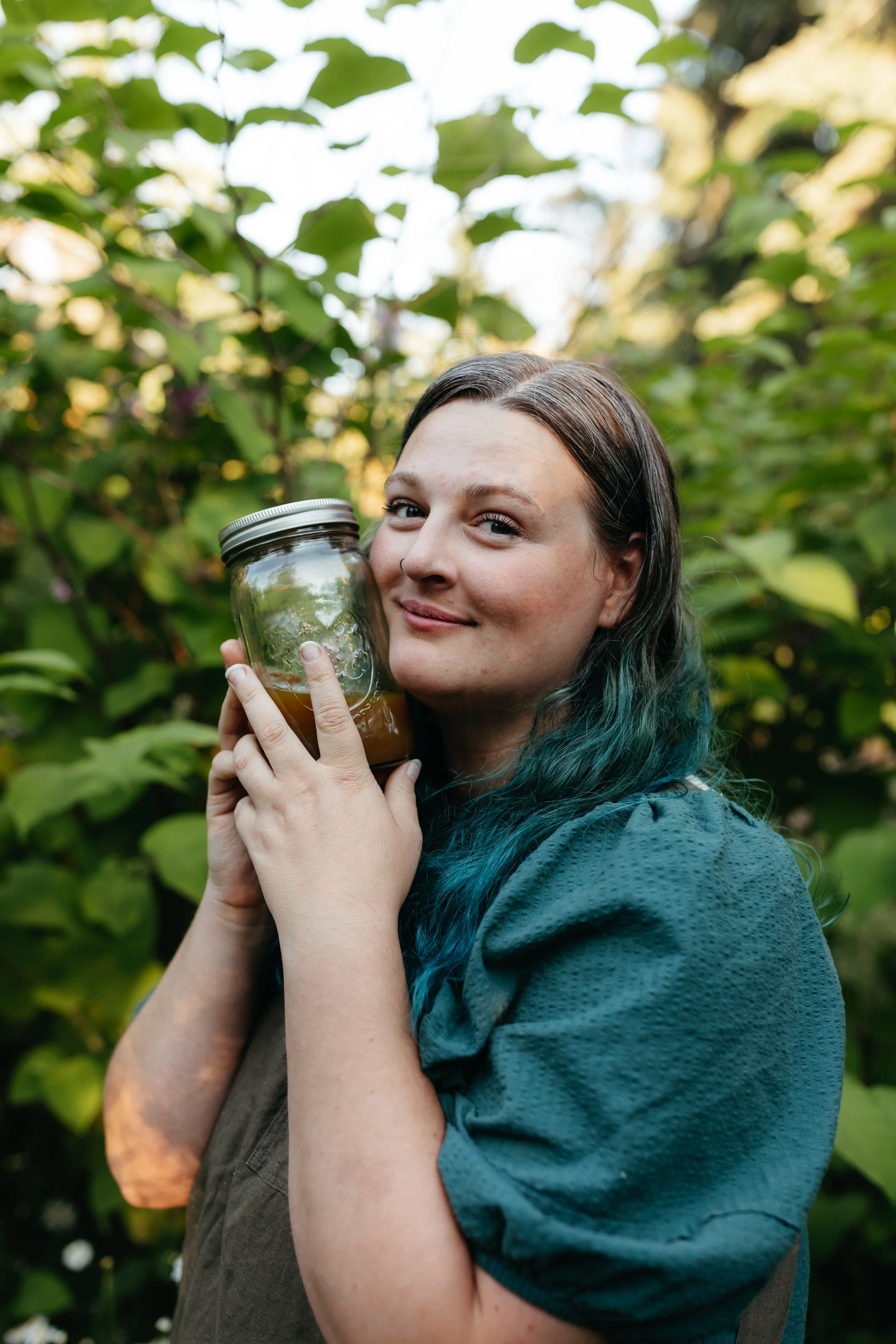 Person holding a mason jar filled with liquid, standing in a garden with lush green foliage.