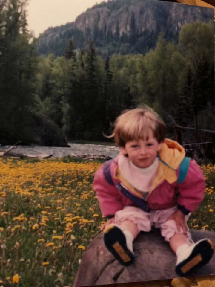 A young child in a colorful jacket sitting on a log in a field of yellow flowers with trees and a mountain in the background.