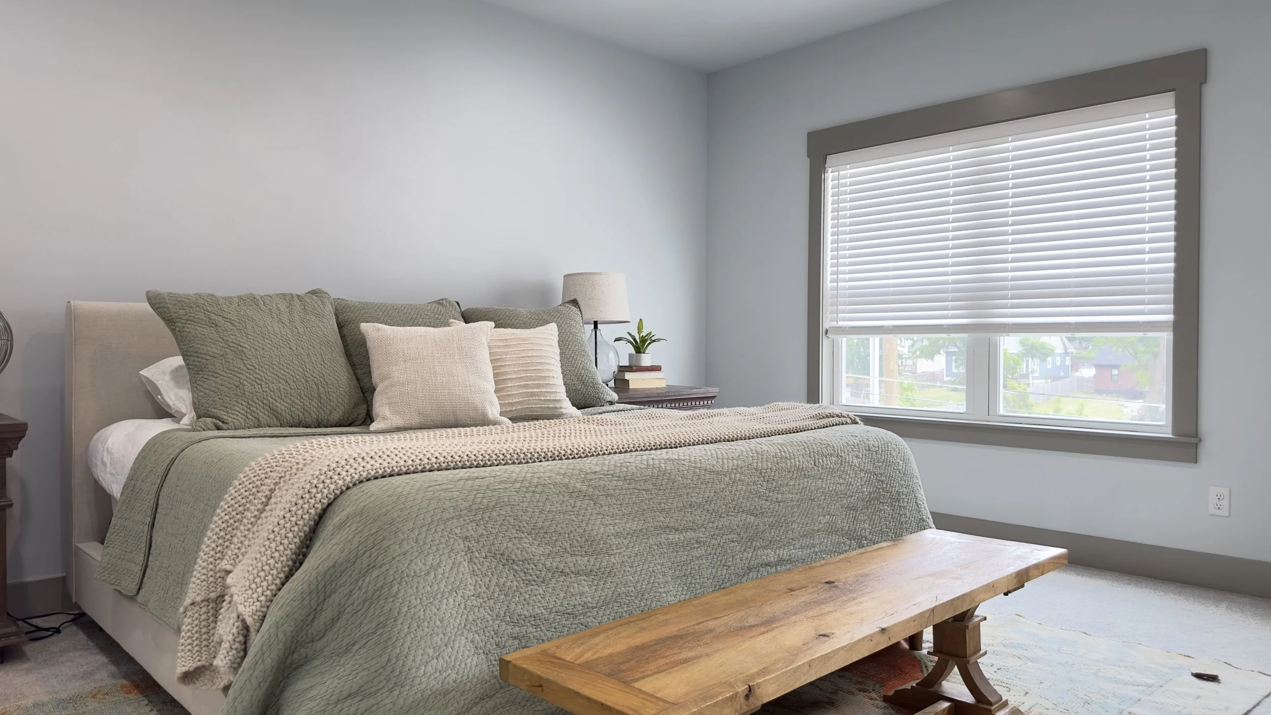 Bedroom with a plush bed, green bedding, wooden bench, nightstand with a lamp, potted plant, and books, large window with blinds.