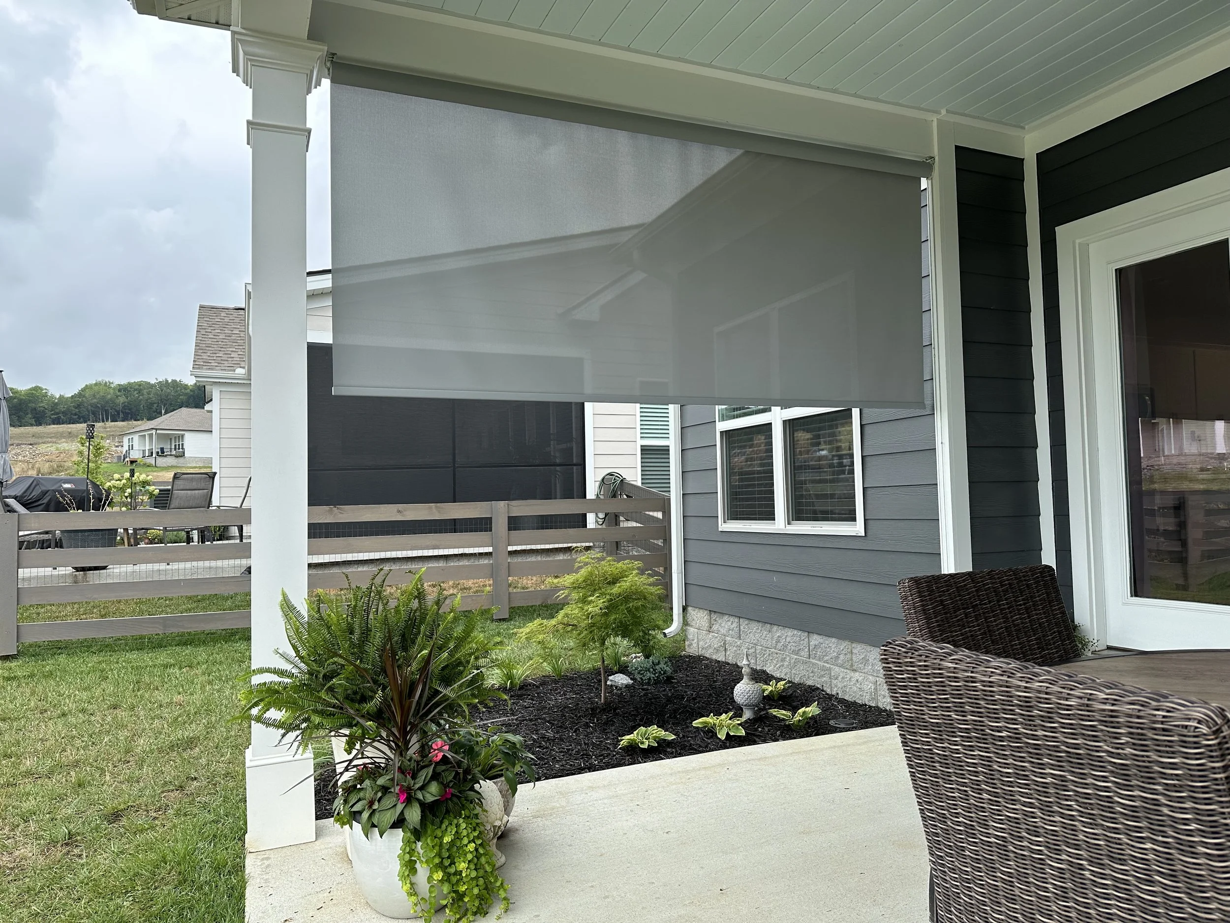 Patio area with woven furniture, a gray sunshade, planters, and mulch landscaping next to a house with gray siding.