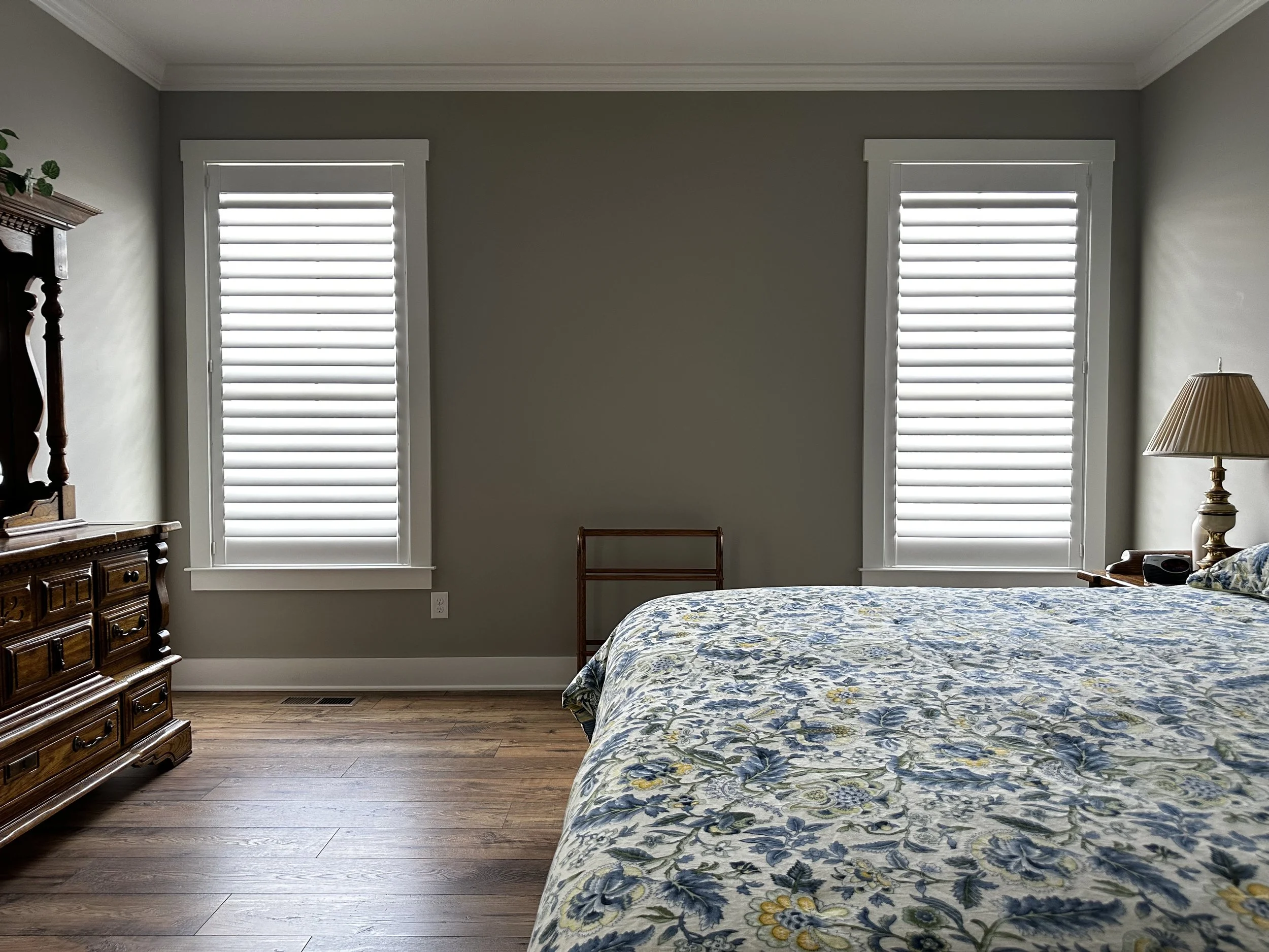 Bedroom with two windows featuring white blinds, a floral-patterned bedspread, a wooden dresser, and a bedside lamp.