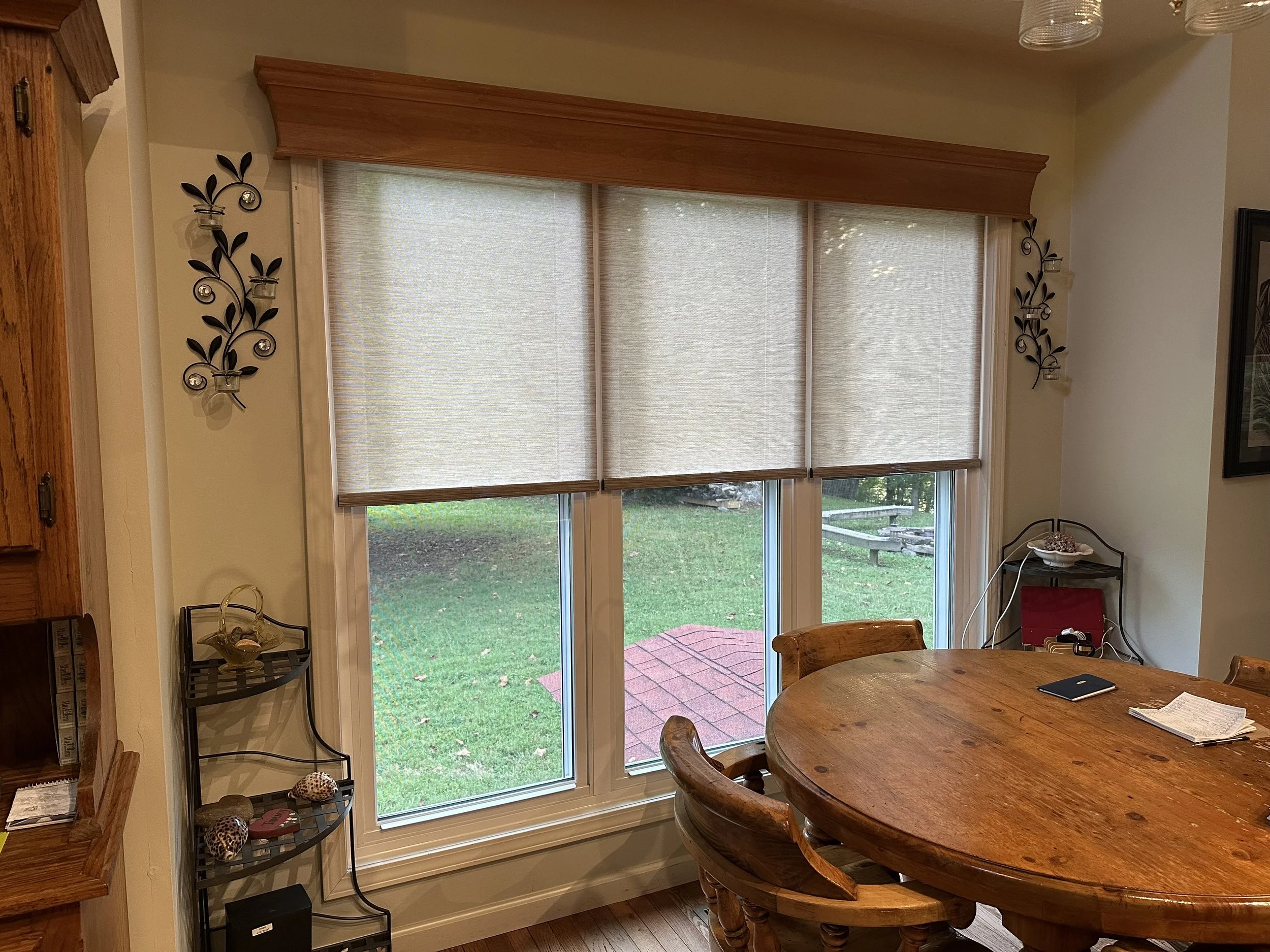 Dining area with wooden round table, wooden chairs, large windows with shades, decorative wall sconces, and small corner shelves with decorative items.