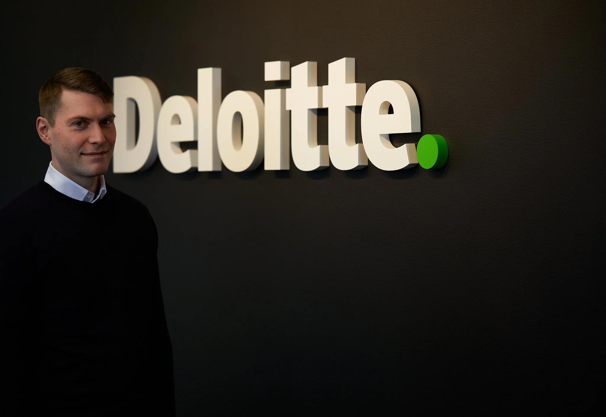 En mann står foran en vegg med Deloitte-logoen. Logoen er i hvitt med en grønn prikk til høyre.
