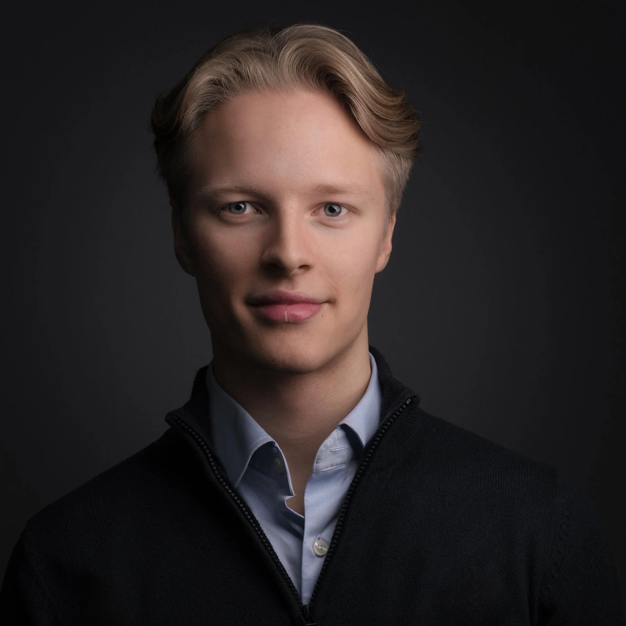 Et portrett av en ung mann med blond hår, blå øyne, iført en lys blå skjorte og en svart genser, mot en mørk bakgrunn.