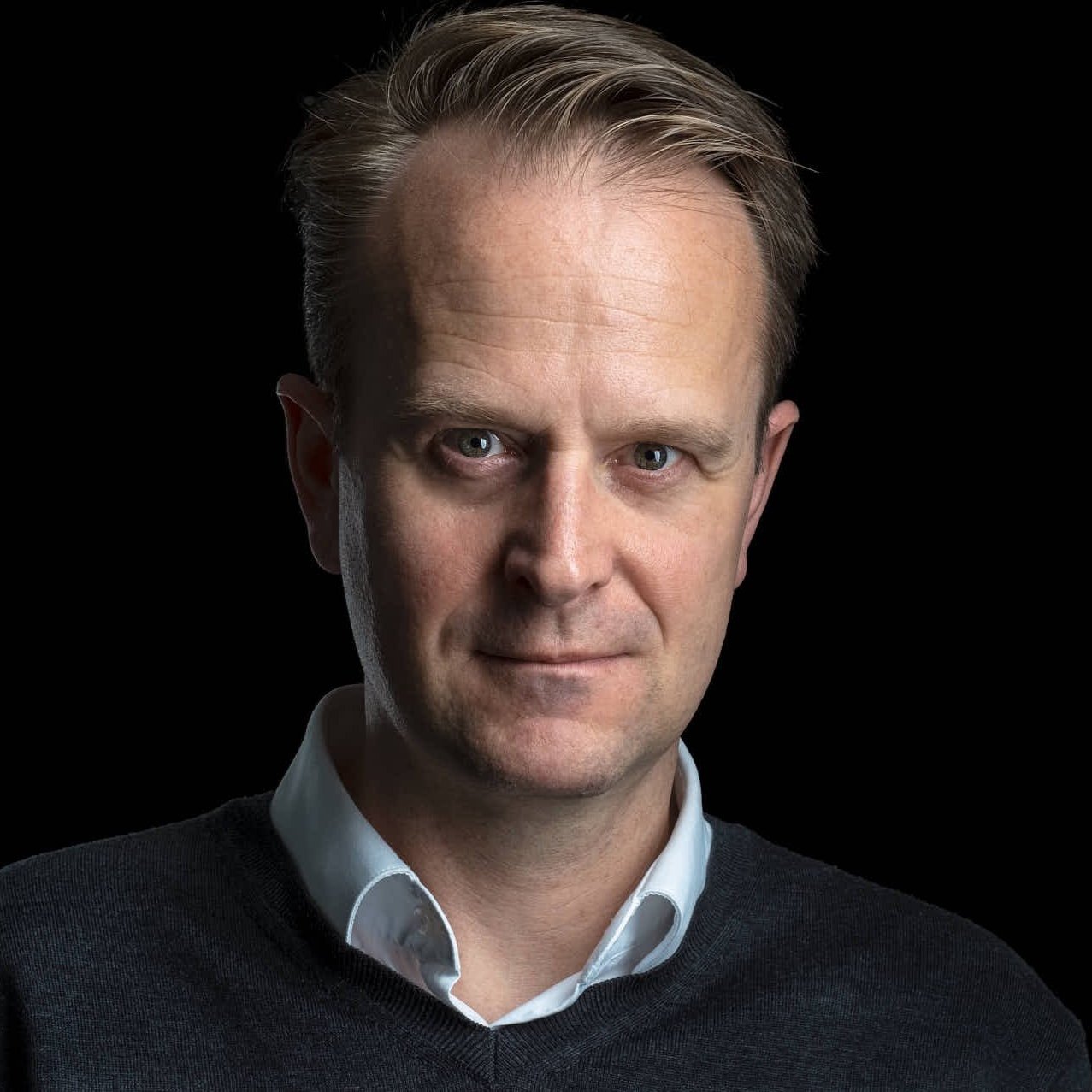 Et portrett av en mann med lyst hår, tydelig på venstre side av bilde, iført hvit skjorte og svart genser, mot en svart bakgrunn.