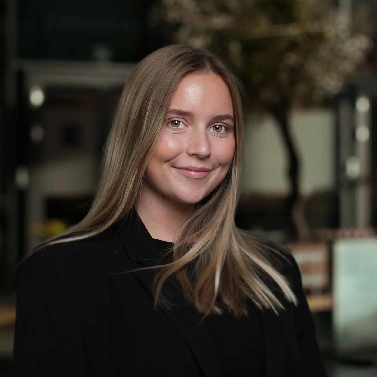 Et nærbilde av en smilende ung kvinne med langt, blond hår, iført svart plagg, foran en utydelig bakgrunn med trær og innendørs elementer.
