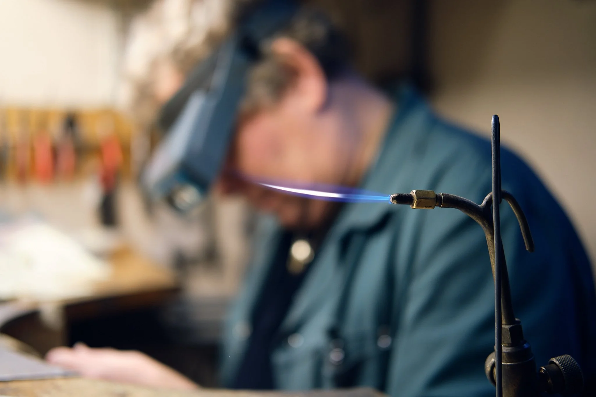 En person i arbeidsgardin og vernebriller som sveiser, med en glødende sveisepistol. Omgivelsene er litt uklare og viser et verksted.