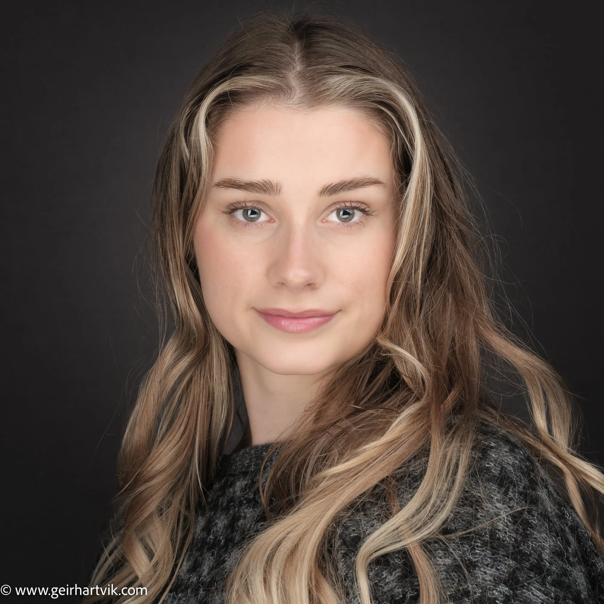 Porträtt av en ung kvinna med långt, blonderat hår och blå ögon, ler mot kameran, mot en mörk bakgrund.