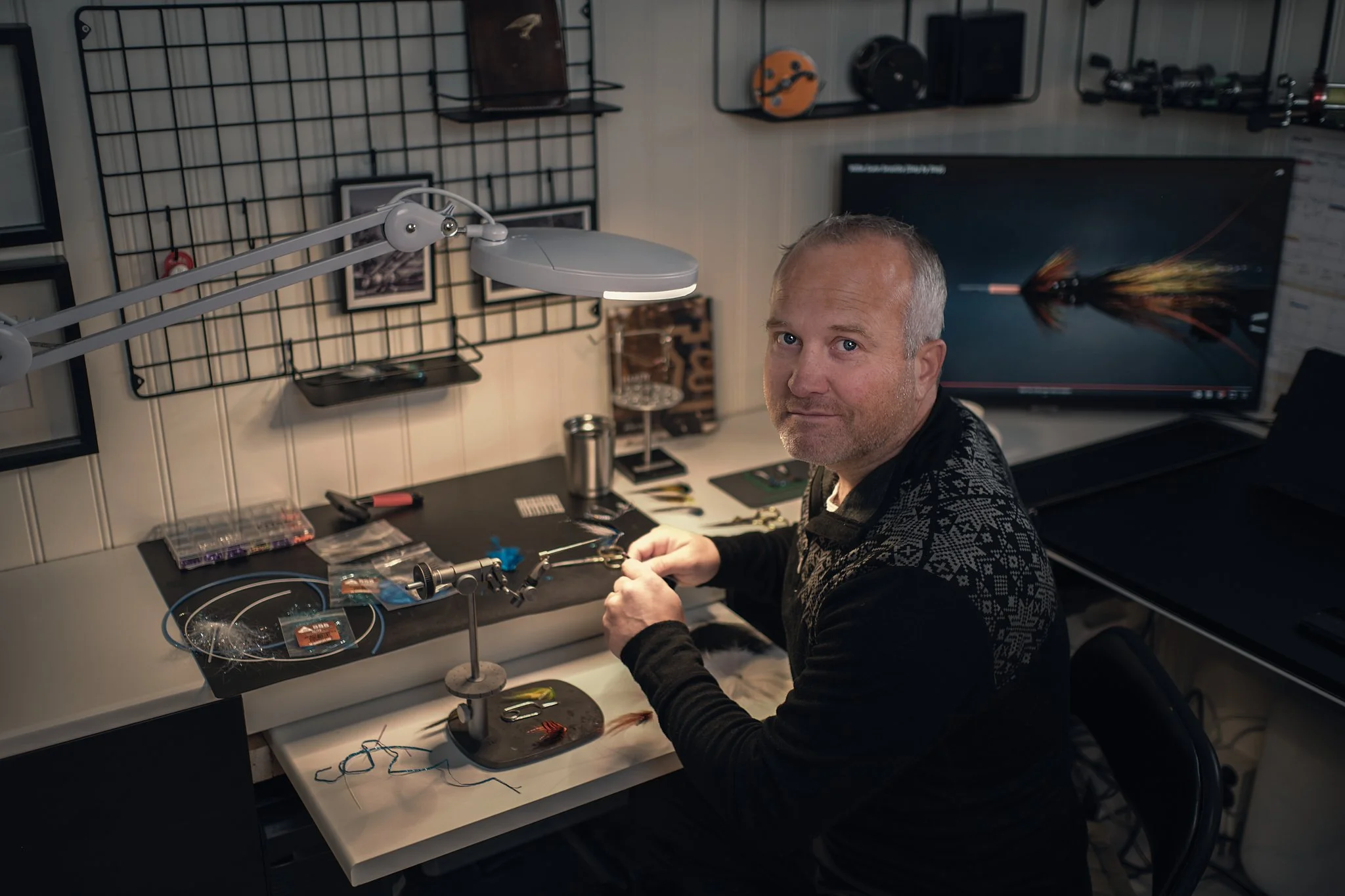 En mann sitter ved et arbeidsbord med verktøy og fiskeassemblingsutstyr, omgitt av fiskerelaterte dekorasjoner og utstyr, med en dataskjerm i bakgrunnen.