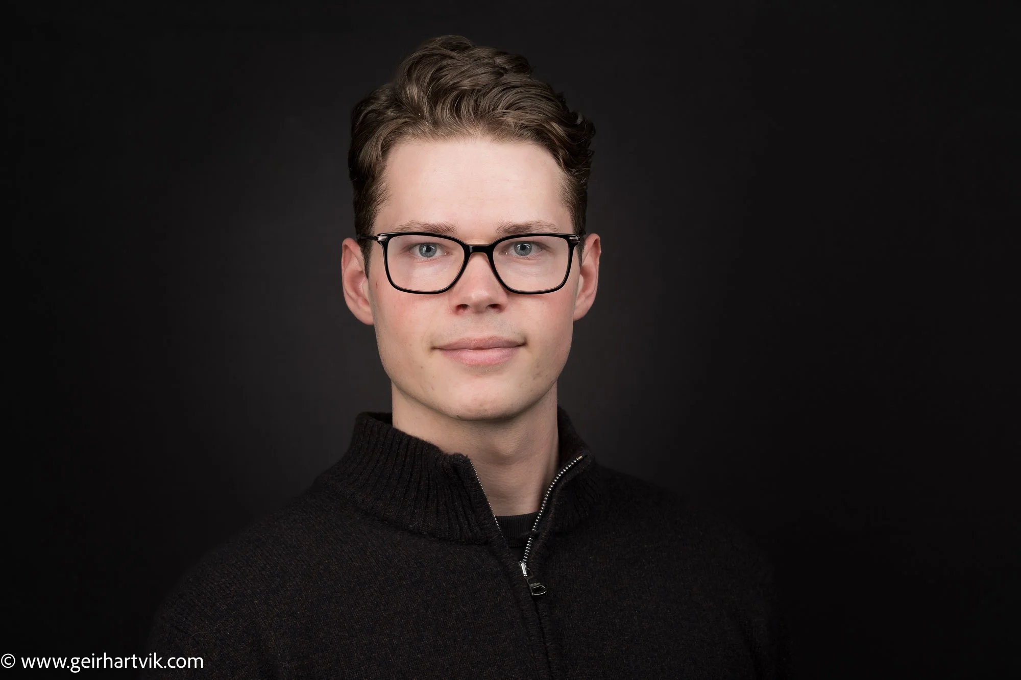 Portrait av en ung mann med brunt hår, iført svarte briller og en svart genser, mot en svart bakgrunn.