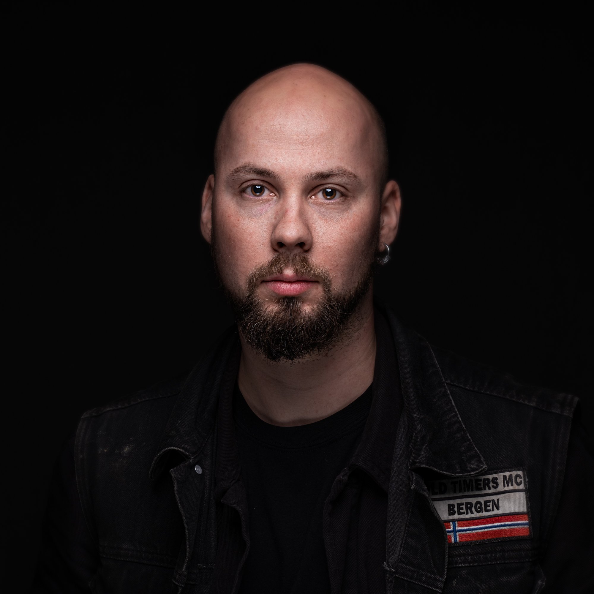 Et nærbilde av en mann med skjegg og skjult hode, iført svart genser og vest som har en patch med teksten 'OLD TIMERS MC BERGEN' og det norske flagget, mot en svart bakgrunn.