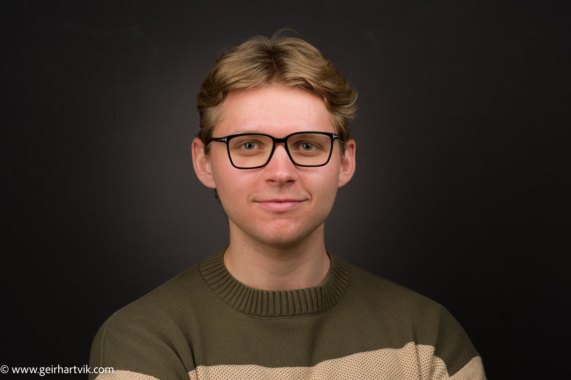 Ung mann med brunt, krøllet hår, iført briller og en grønn og beige strikkegenser, smilende foran en svart bakgrunn.