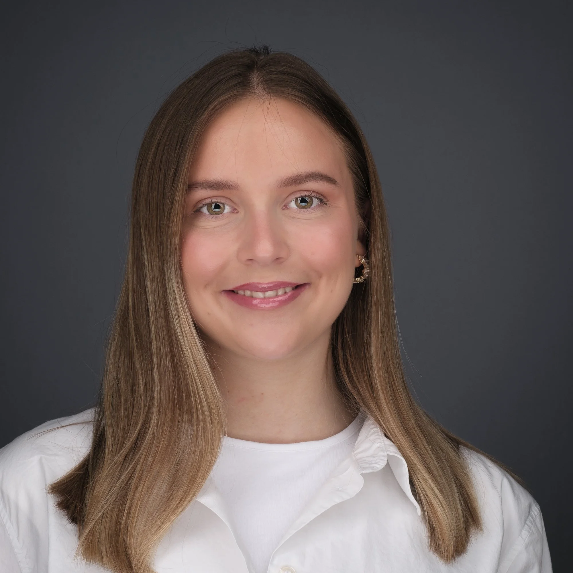 Portrett av en smilende kvinne med langt, brunt hår og epleformede øyne, iført hvit bluse, mot en mørk grå bakgrunn.