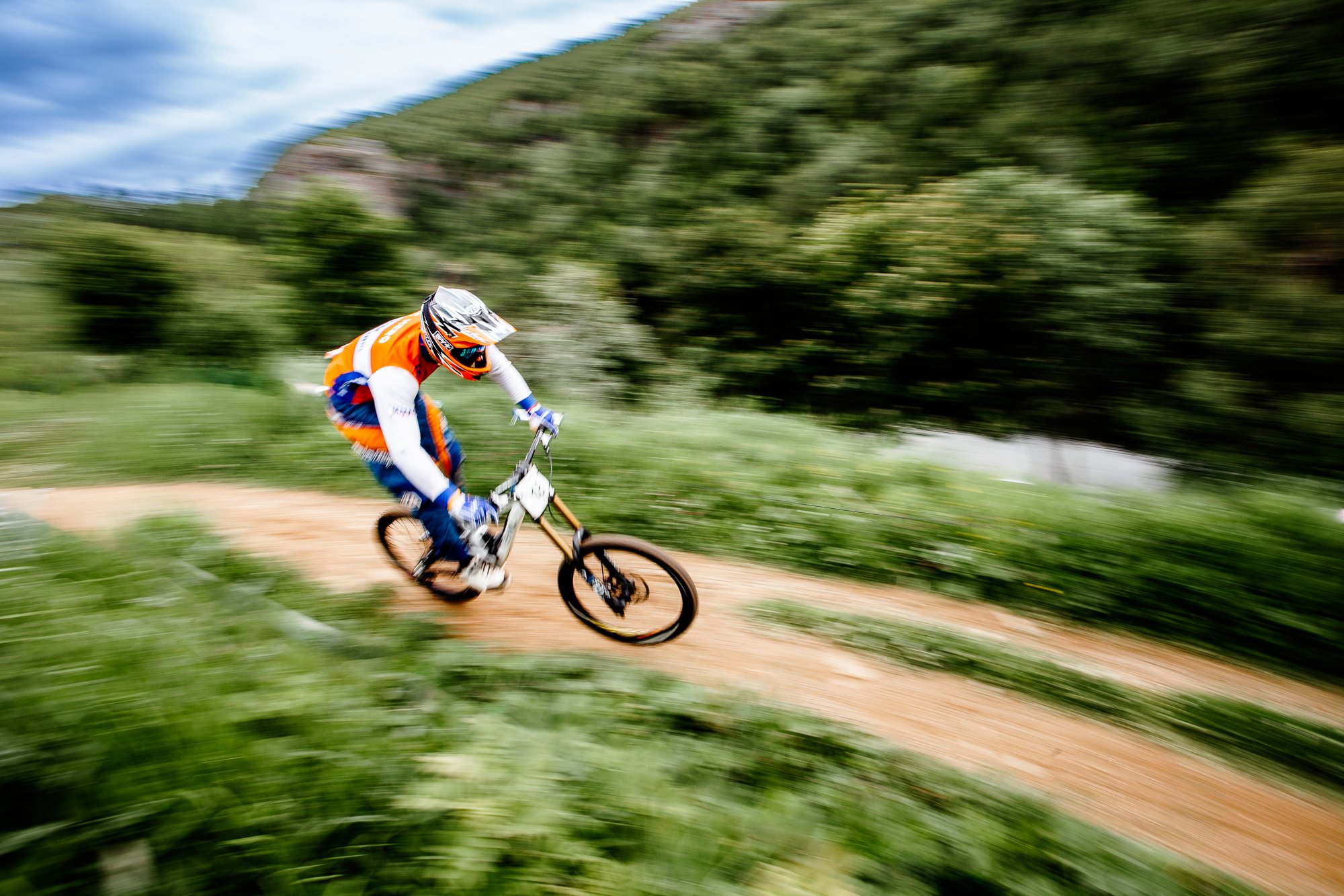 Sykkelfører i full fart på en grusvei, med alvorlig fokus, omgitt av grønt landskap og fjell i bakgrunnen.