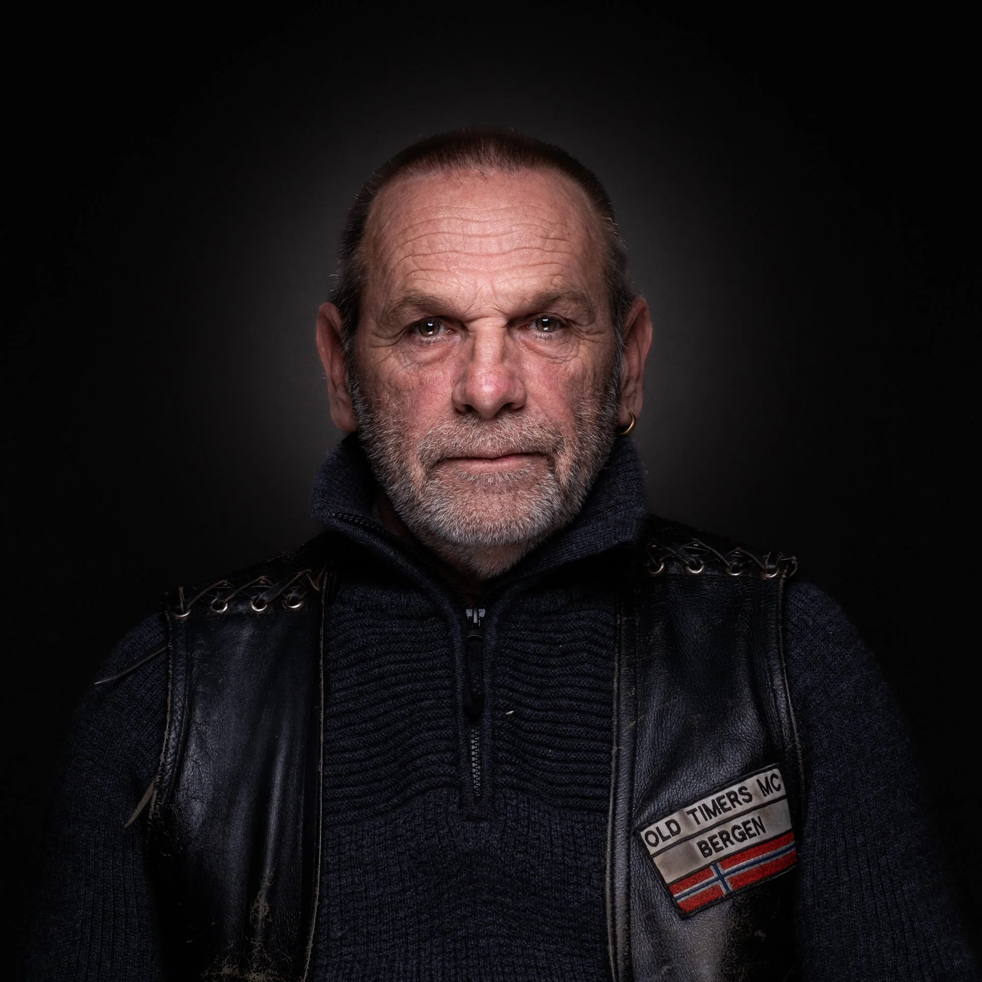 Portrett av en eldre mann med kort, grått hår og skjegg, iført svart klær og skinnvest med 