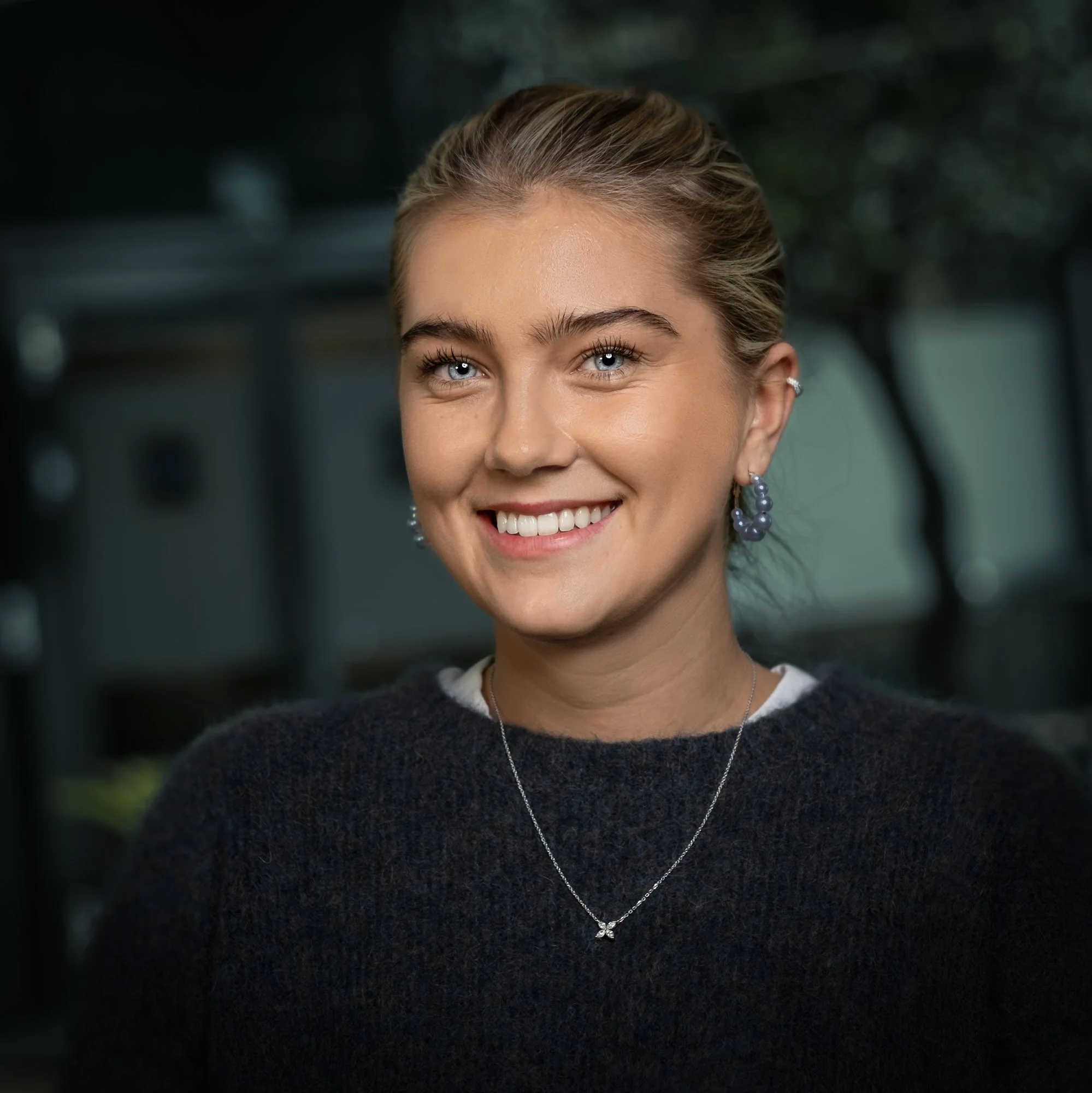 Et smilende ungt kvinne med blondt hår og blå øyne, iført sølvøreringer, et sølvhalskjede med en liten anheng, og en mørk sweater, ute i en park eller hage.