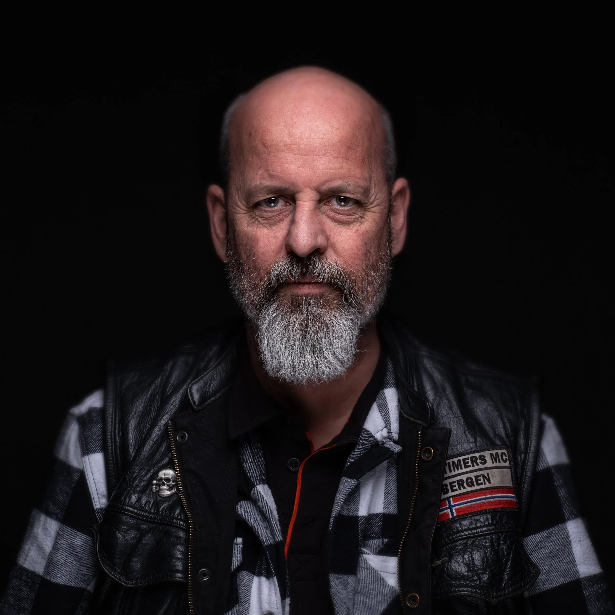 Portrett av en mann med grått skjegg, iført svart og hvitt rutete skjorte og skinnvest med patcher, mot en svart bakgrunn.