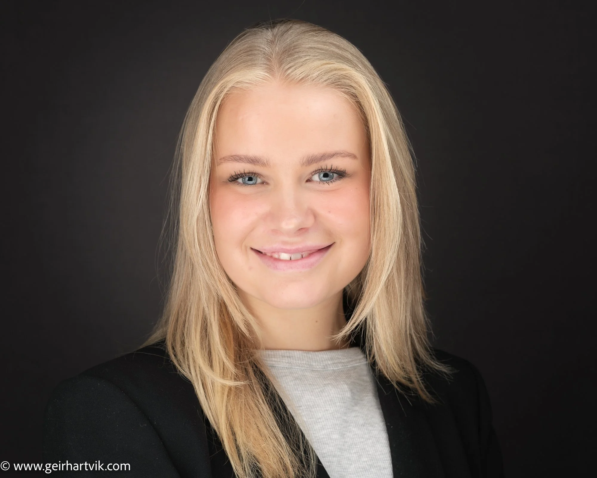 Portrett av et smilende kvinnekort med blondt hår, lys hud, blå øyne, svart blazer og lysegenser mot svart bakgrunn.