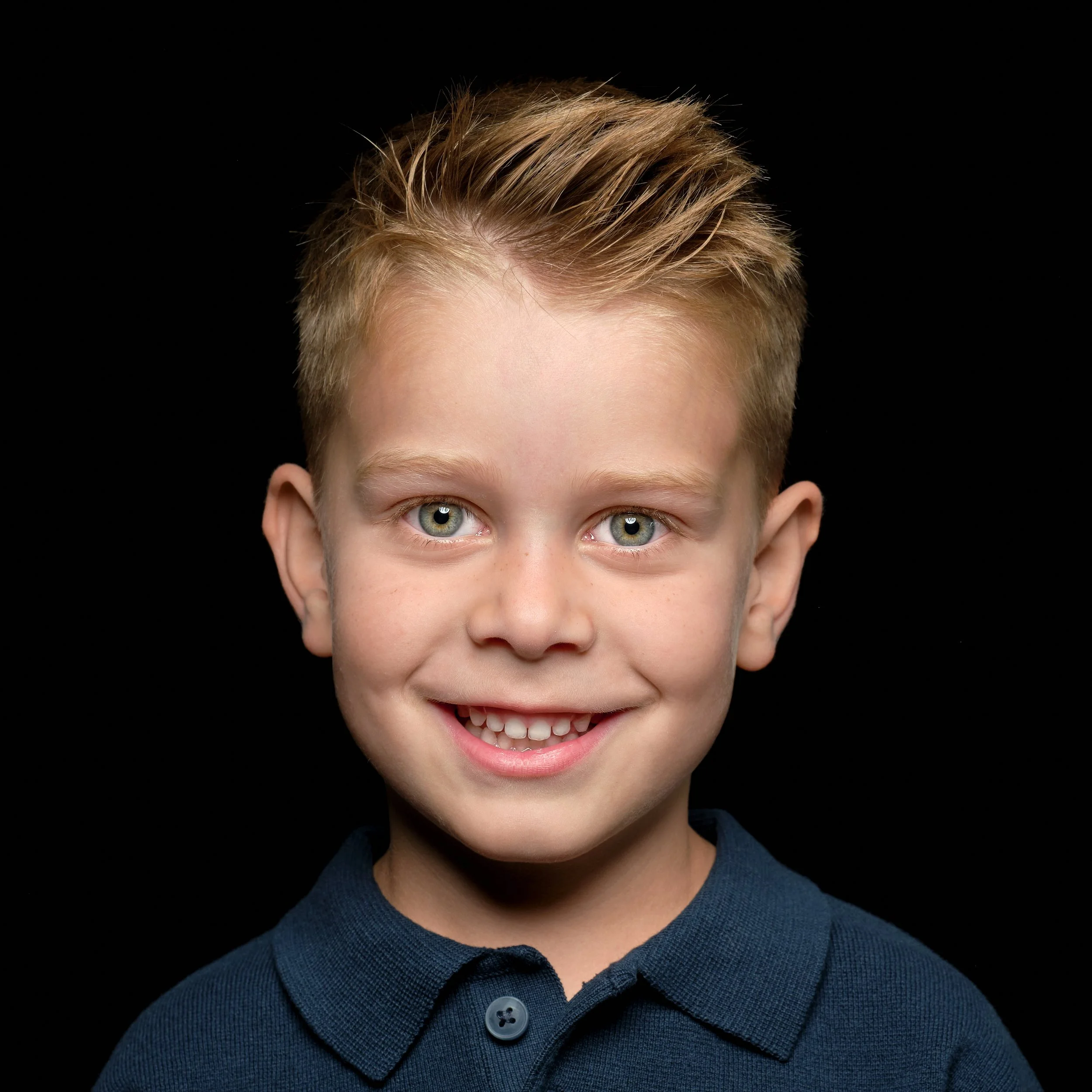 Et smilende ungt barn med lyst, kort hår, iført en mørkeblå polo mot en svart bakgrunn.