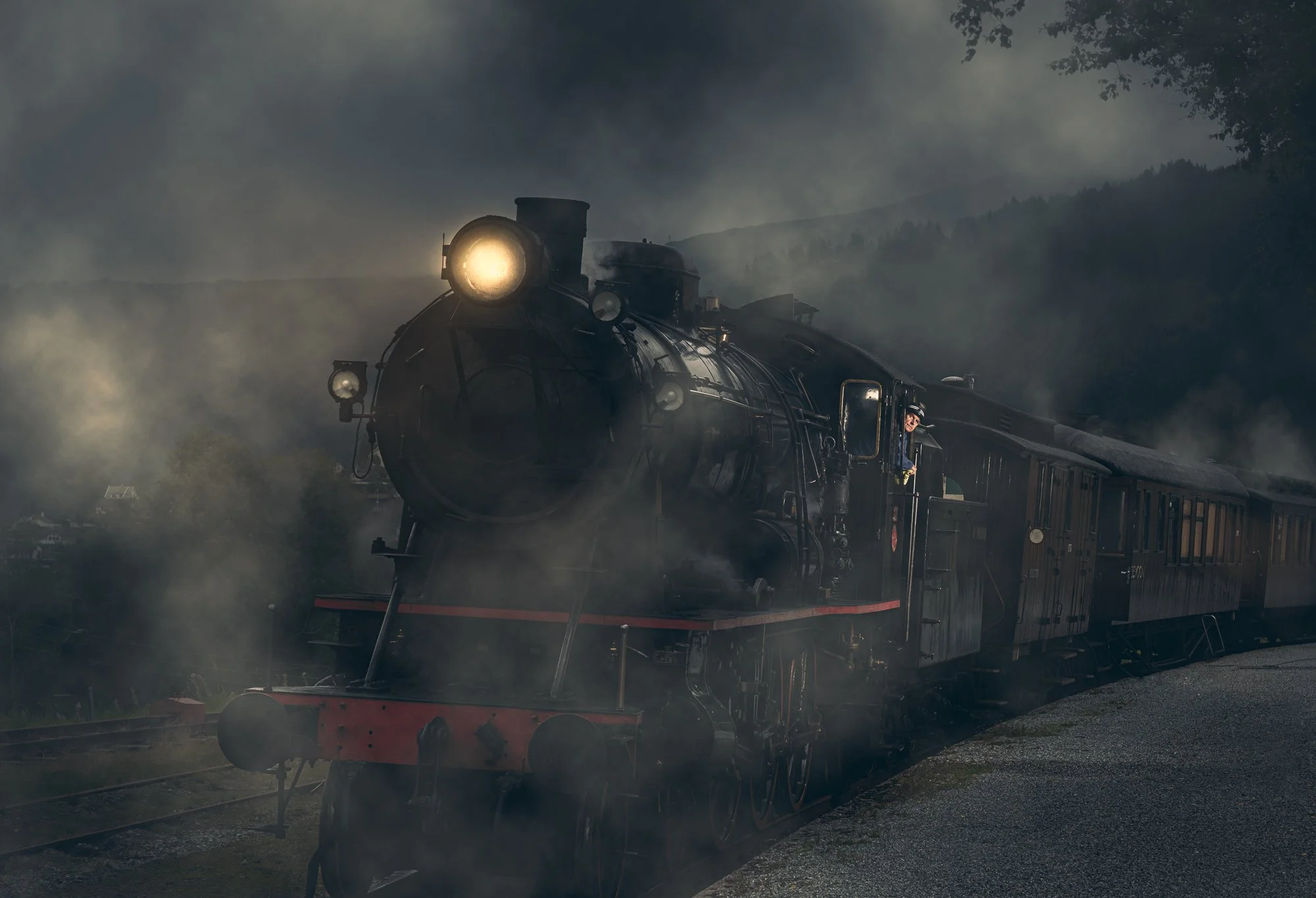 En svart damp- og kulltype lokomotiv som kjører ut av tåke på en stasjon, med fjell og trær i bakgrunnen.